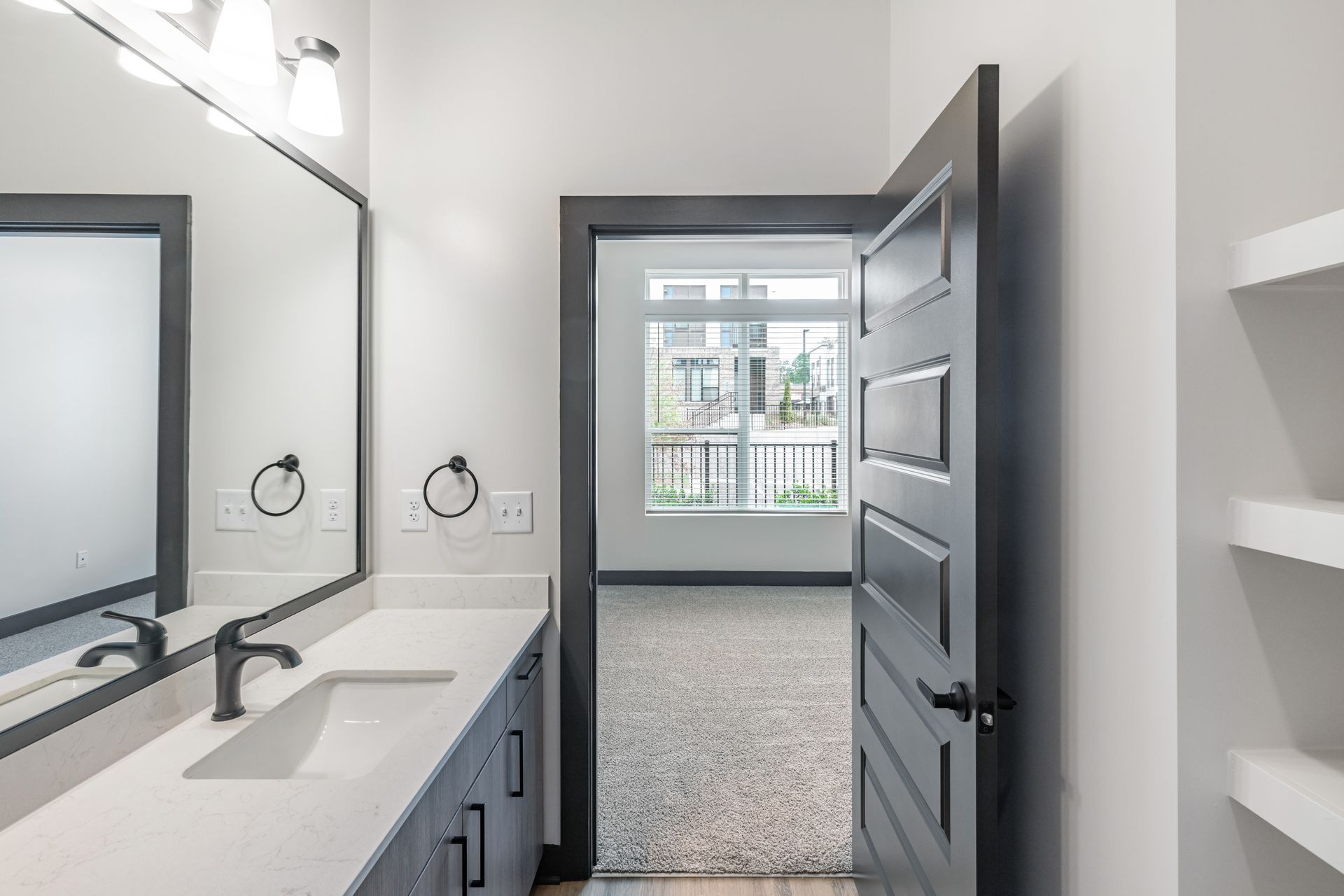 Modern bathroom with double vanity, large mirror, open door, and window to a bedroom beyond at Vela Park apartments in East Lake, Atlanta, GA.