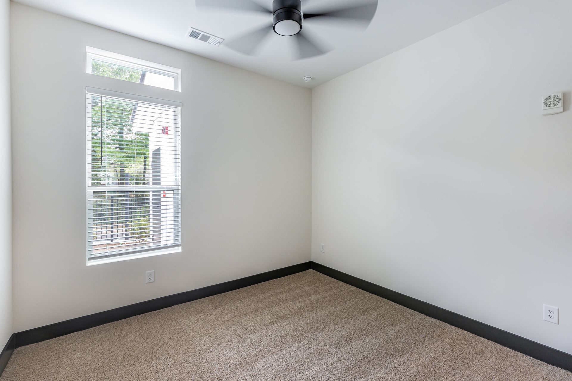 Empty bedroom with white walls, carpeted floor, ceiling fan, and a window with blinds at Vela Park apartments in East Lake, Atlanta, GA.