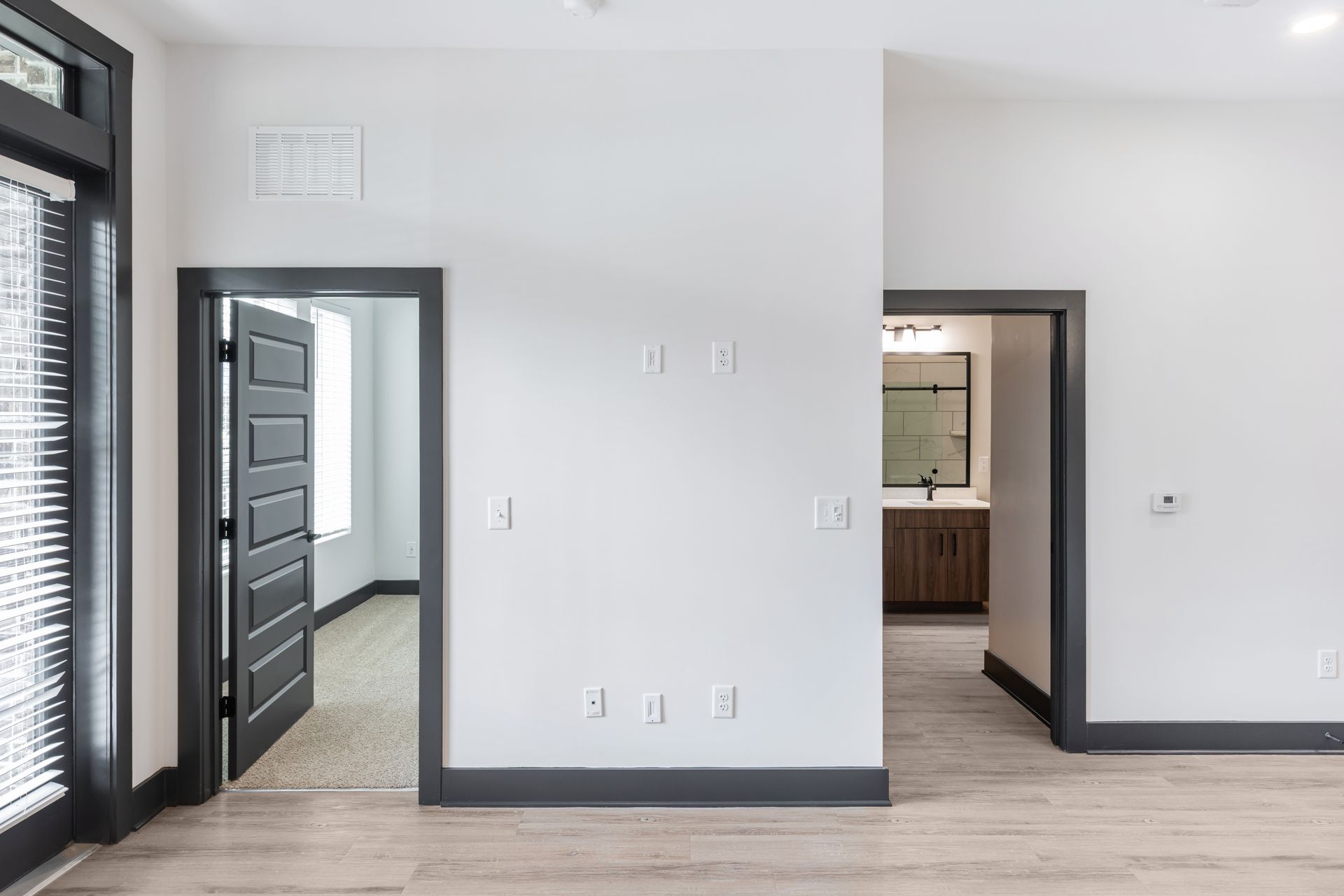 Empty modern room with white walls, gray trim, wood floor, and two doorways to a hallway and kitchen at Vela Park apartments in East Lake, Atlanta, GA.