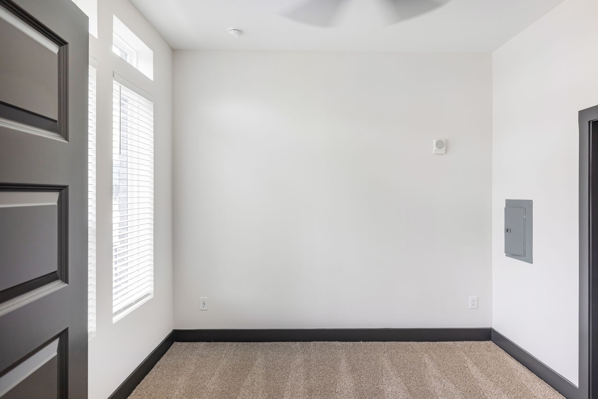 Empty white room with carpeted floor, black doorframe, window blinds, and wall panel on the right at Vela Park apartments in East Lake, Atlanta, GA.
