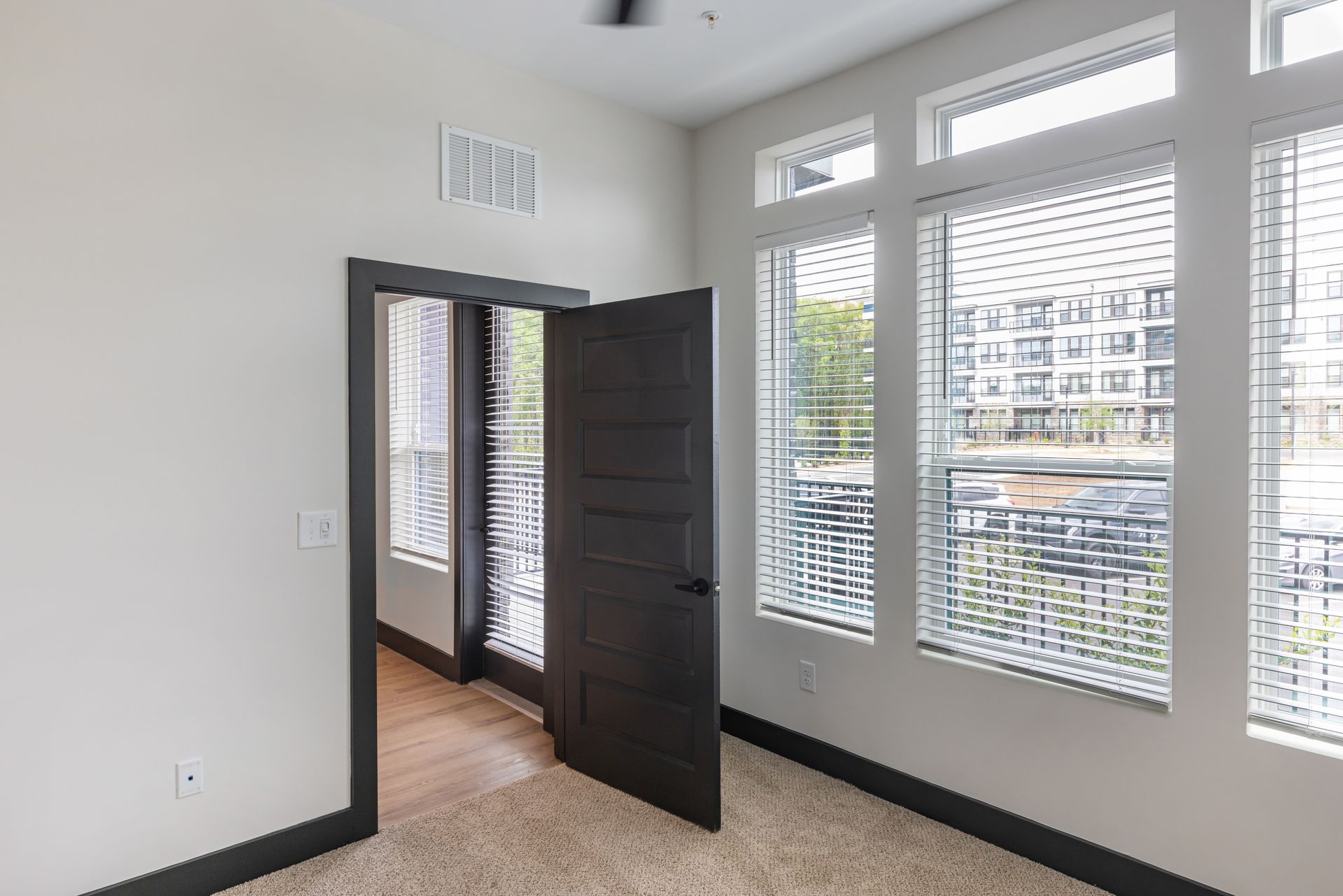 Bright empty room with large windows, white walls, and an open dark door onto a hallway at Vela Park apartments in East Lake, Atlanta, GA.