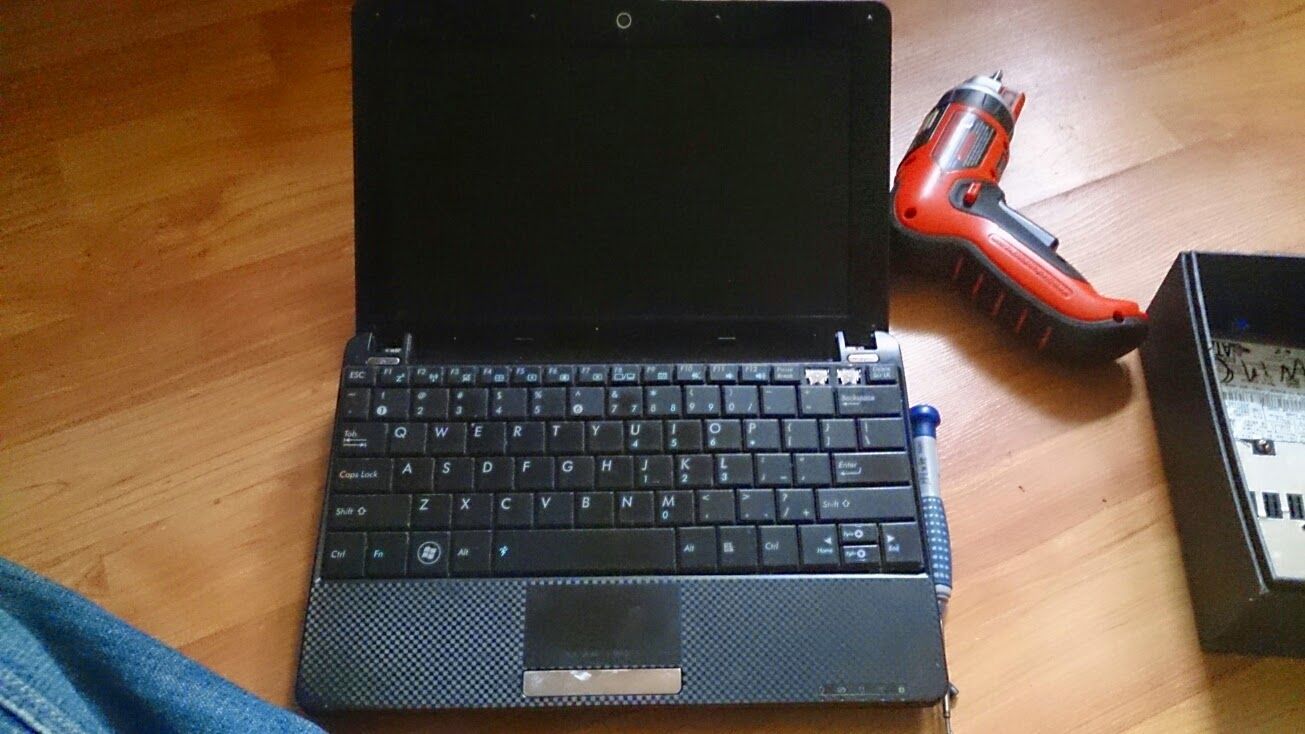 Laptop with keyboard open, red power drill and case on wooden floor.