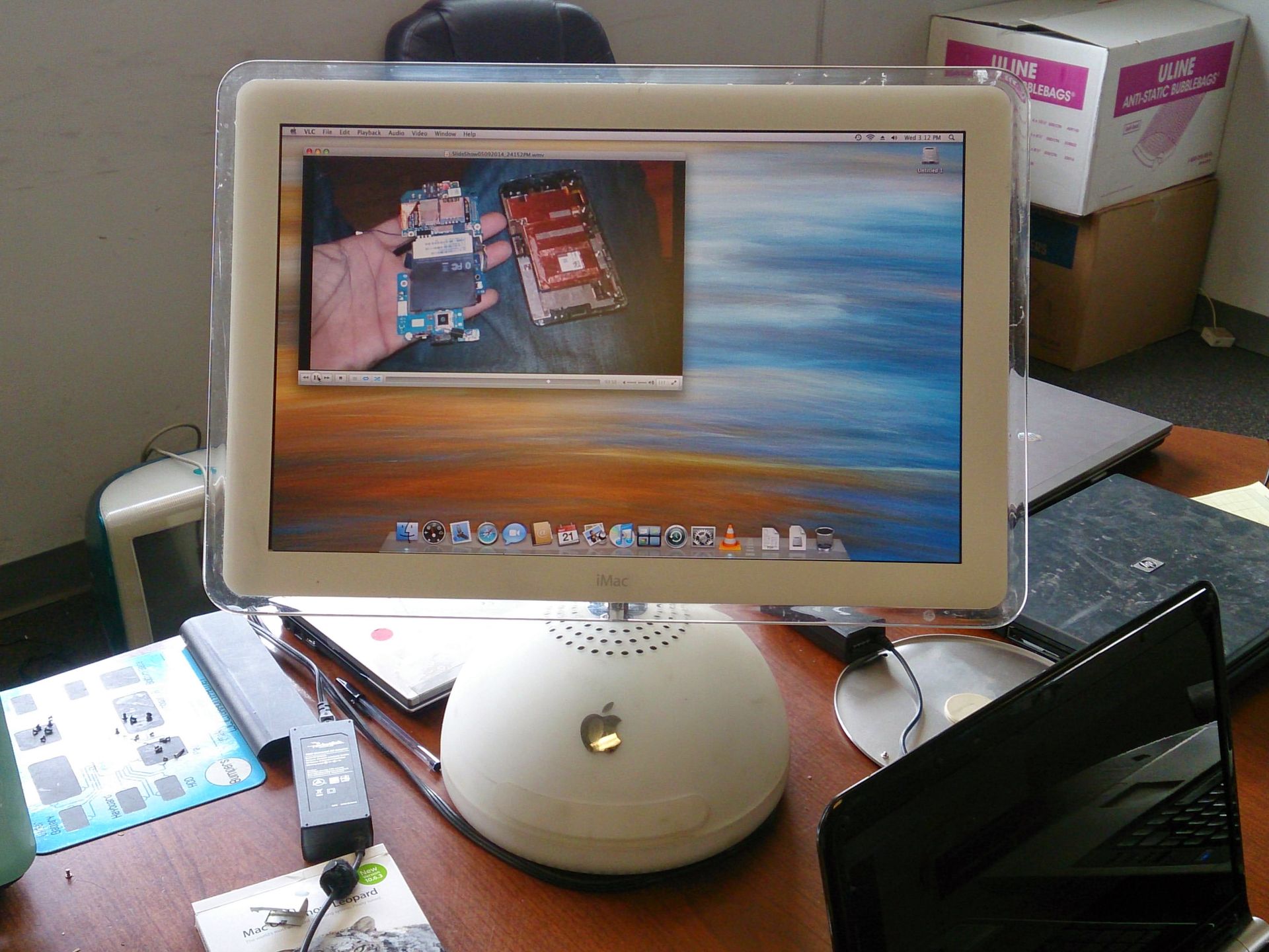 An iMac computer with a screen displaying a photo of circuit board components. Other laptops and objects on a desk.