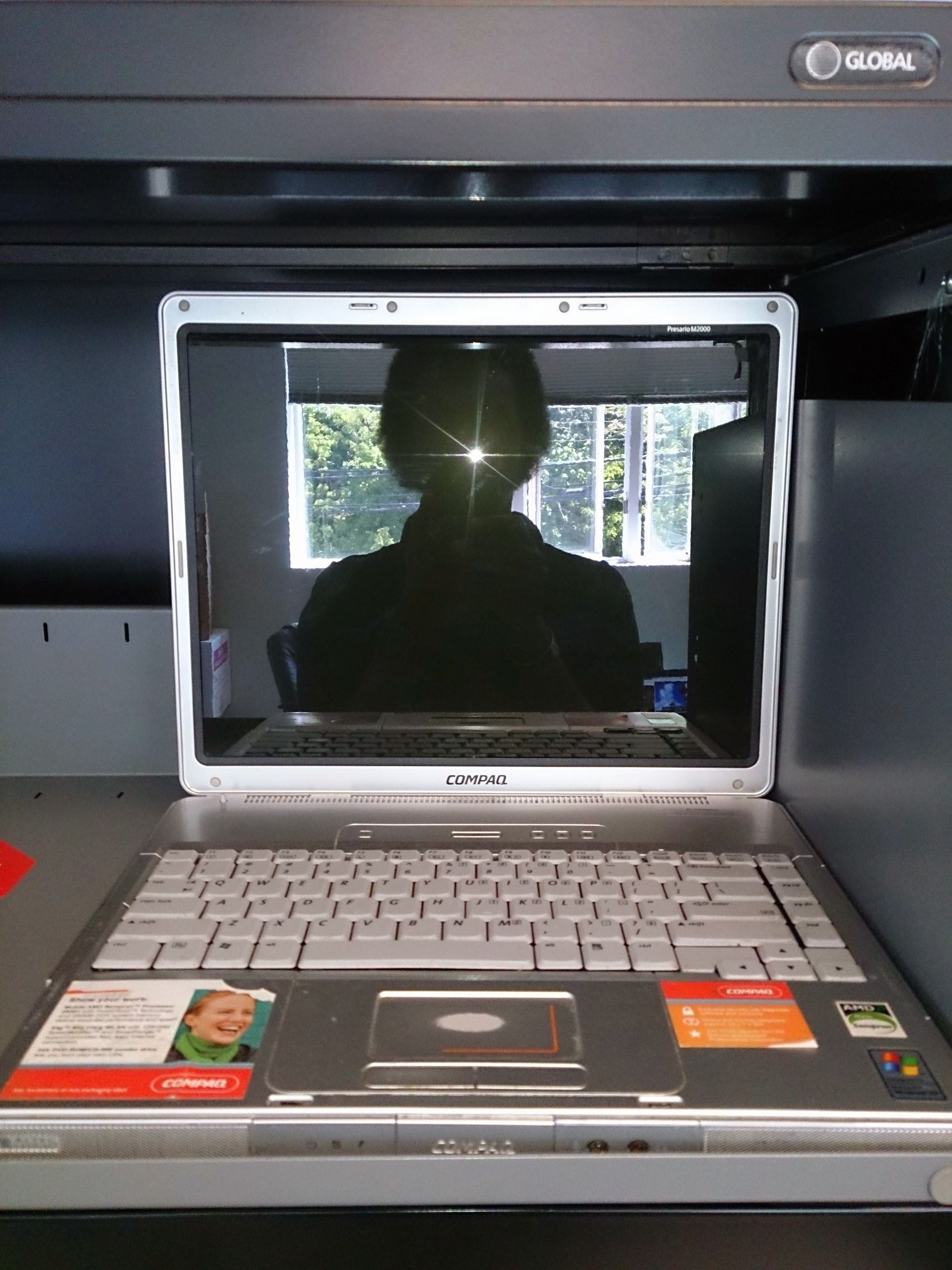 Silver laptop open, reflecting a person and a bright window. Keyboard visible, resting on a shelf.