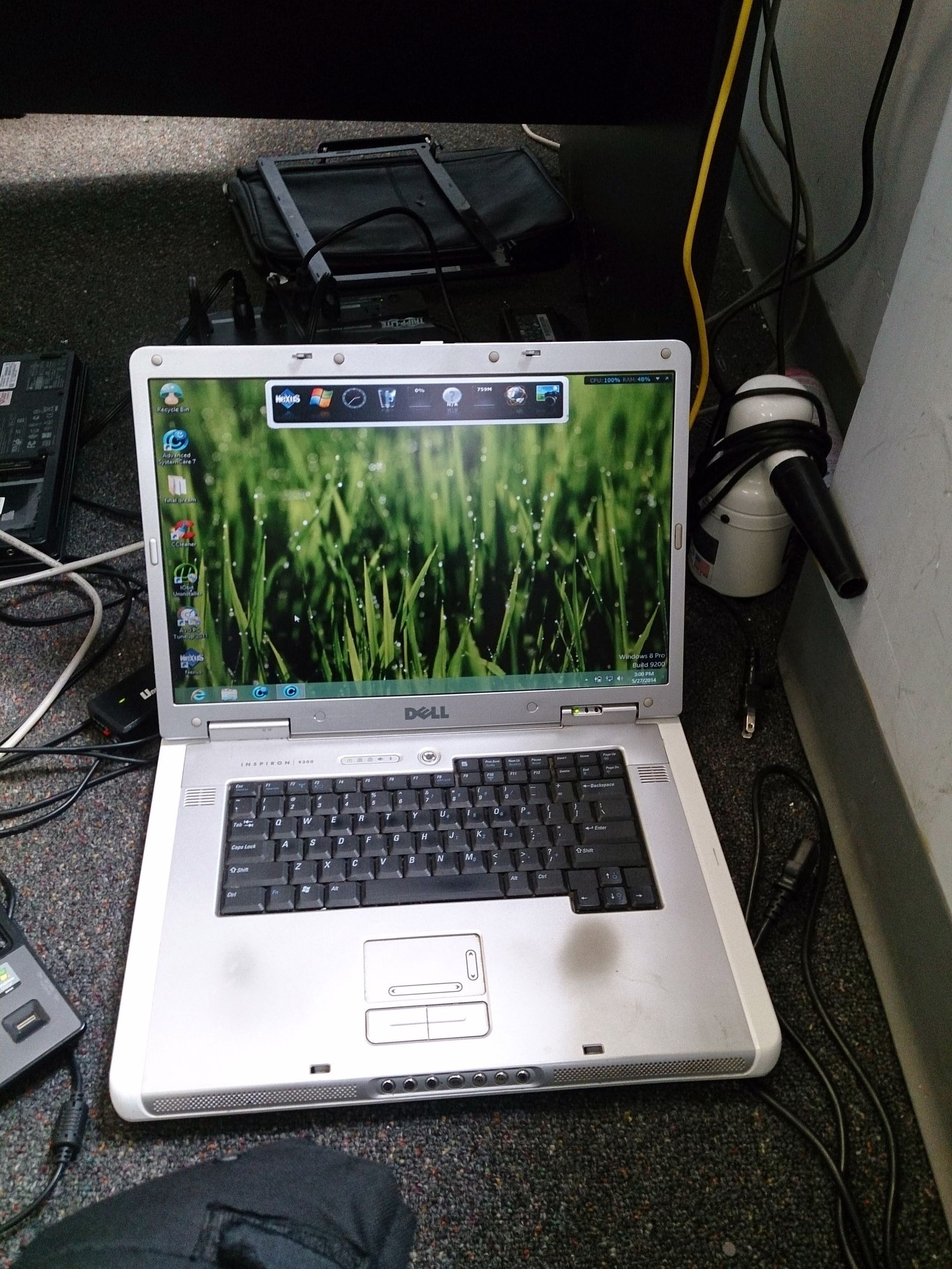 Silver Dell laptop on a dark floor, open with a green grass wallpaper. Cables and equipment surround it.