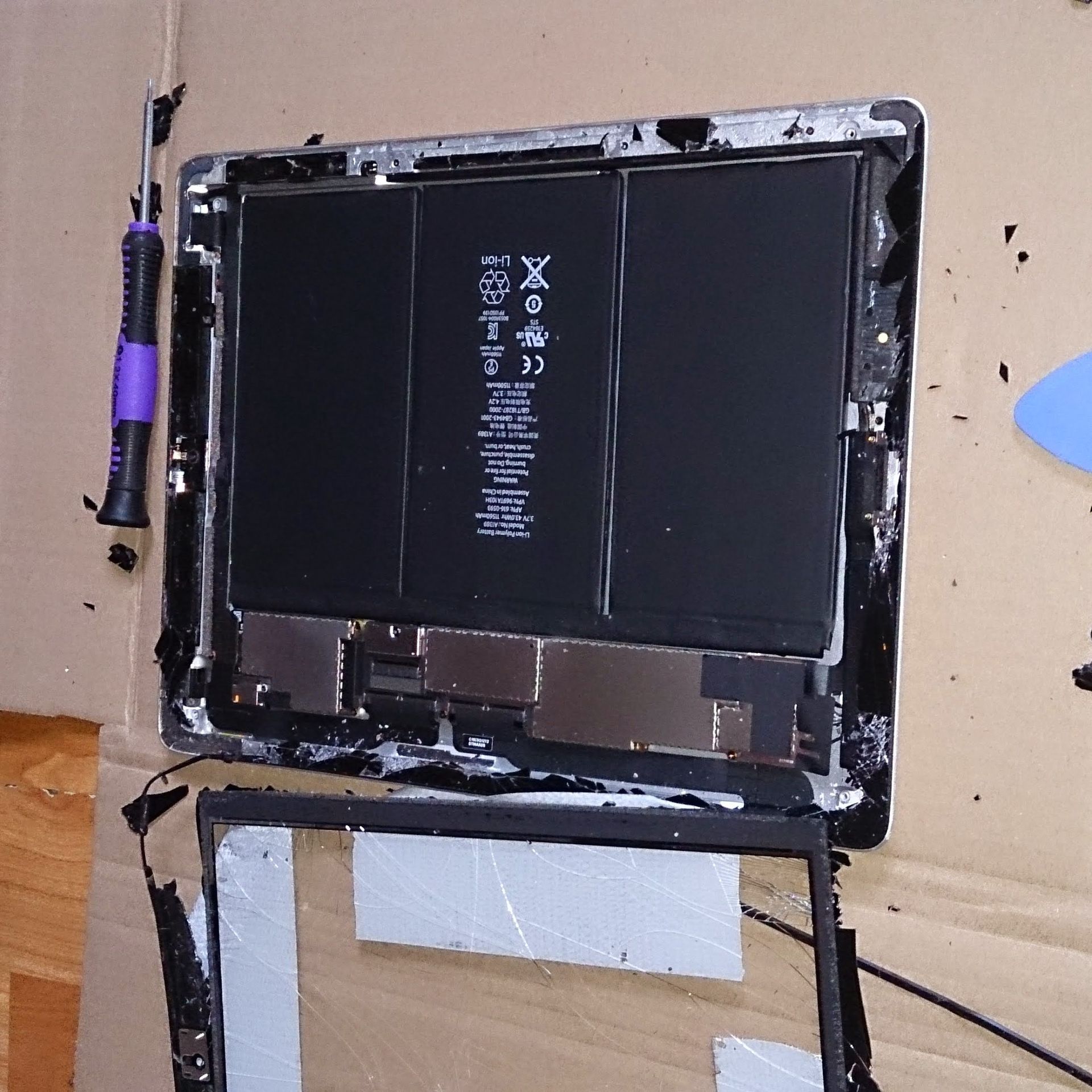 Disassembled tablet with exposed battery and internal components, lying on a cardboard surface.