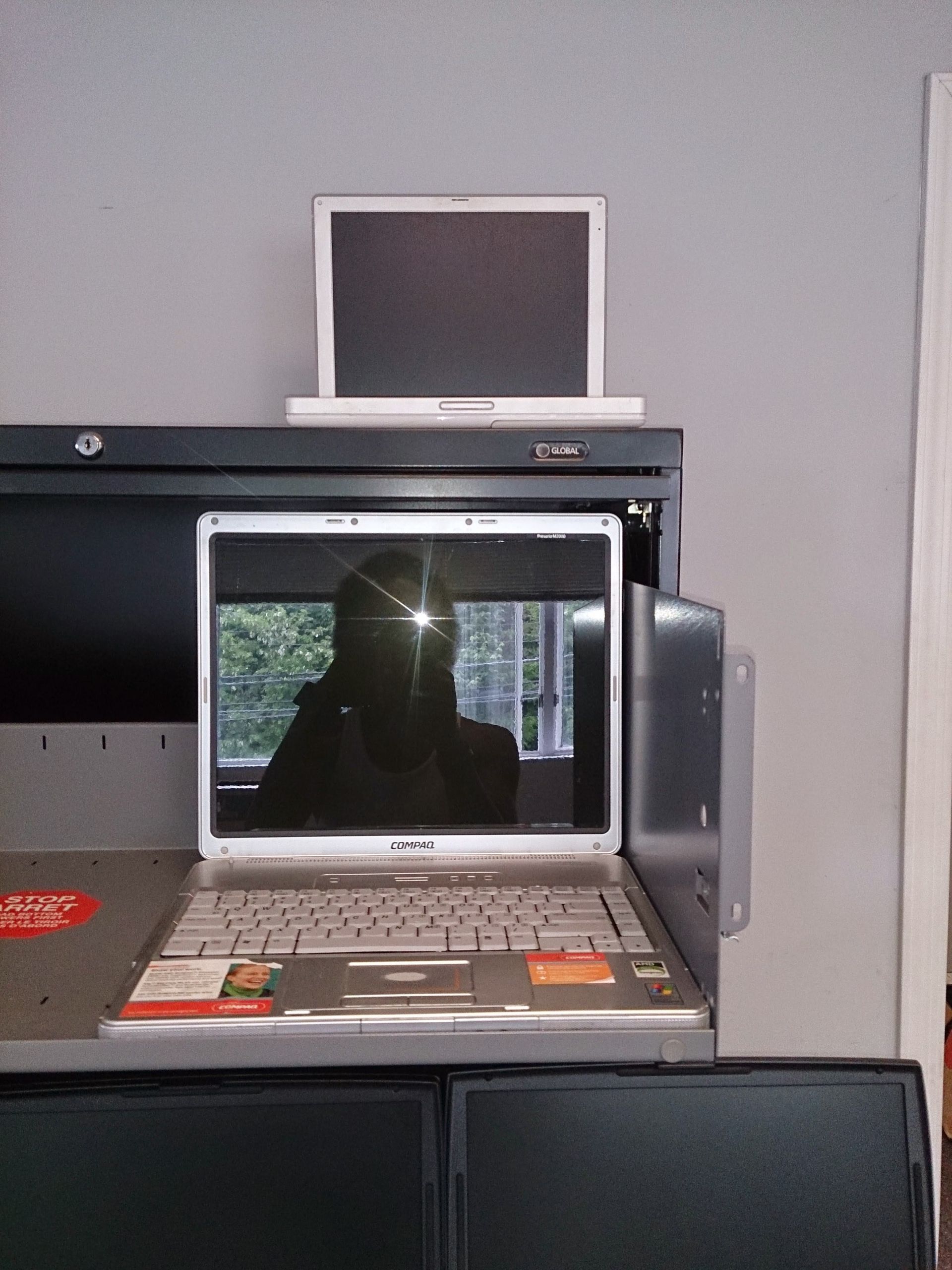 Two laptops stacked on a shelf. One open, reflecting a window. The other closed on top.