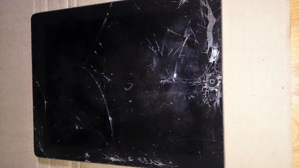 Damaged black tablet screen with extensive cracks, resting on a cardboard surface.