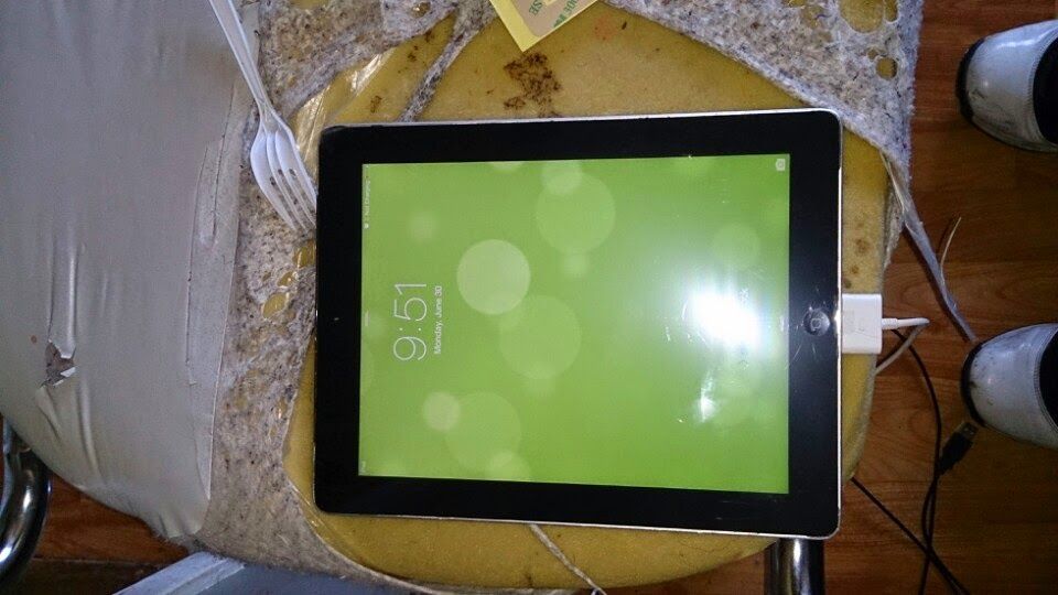 IPad with green screen displaying 9:51, resting on a worn, yellow cushion; charging cable connected.