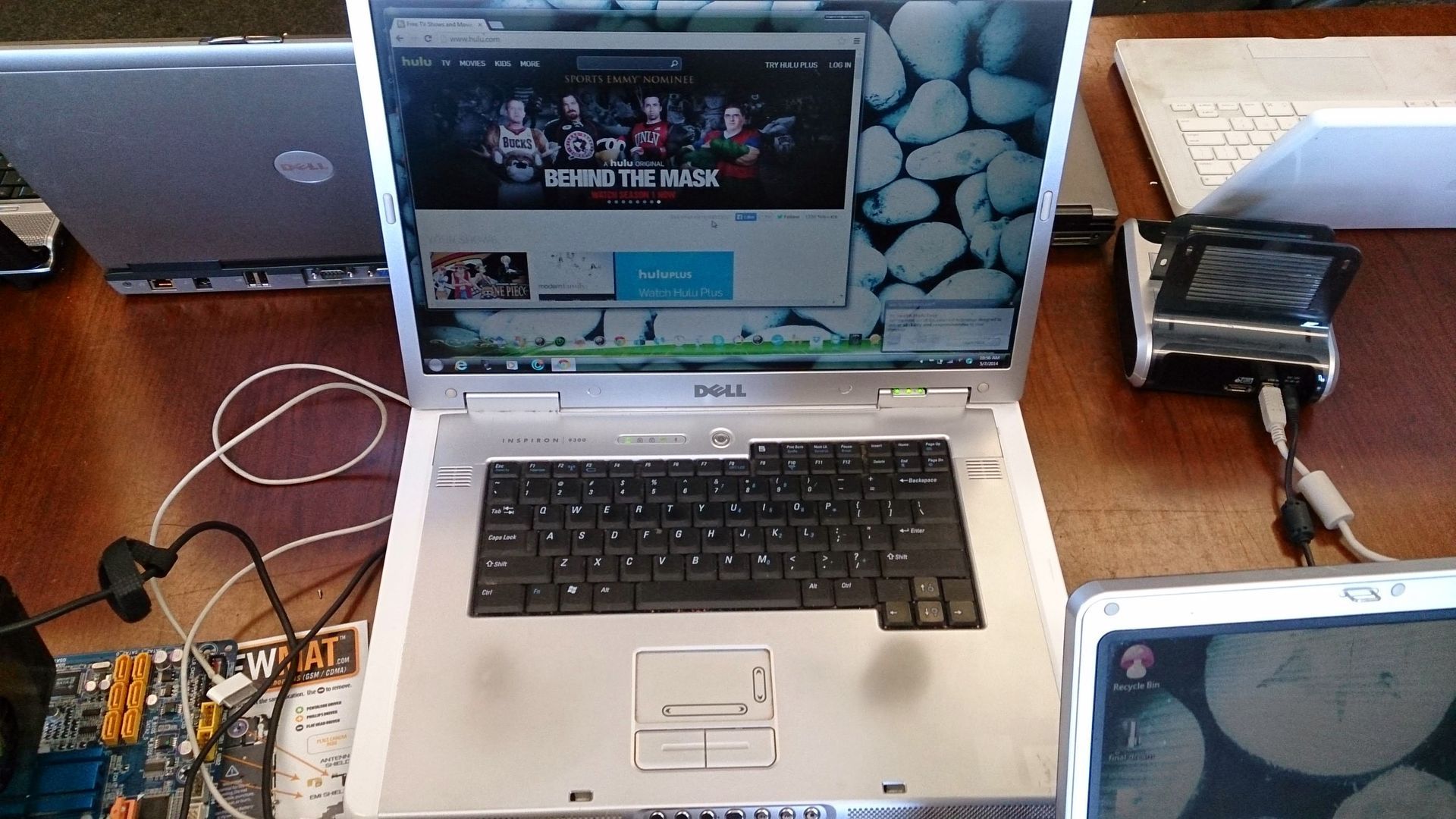 A laptop with a video playing on screen, surrounded by other laptops and electronics on a wooden surface.