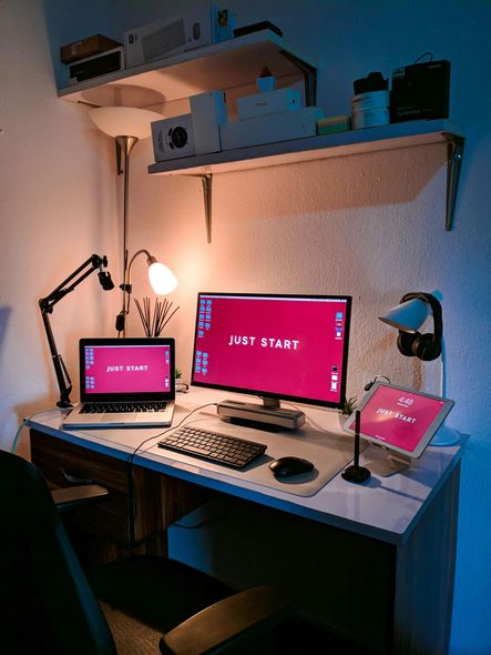 Desk with two monitors, keyboard, mouse, and other office supplies lit by desk lamp and shelf lighting.