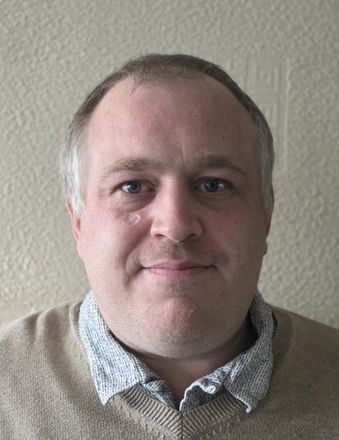 Man with light skin, light brown sweater, and patterned shirt.
