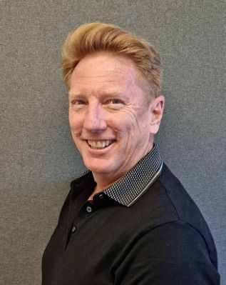 Man with red hair and a black shirt smiling in front of a gray wall.