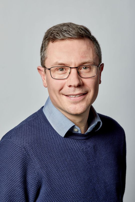 A man in glasses smiles at the camera. He wears a blue sweater over a blue and white checkered shirt.