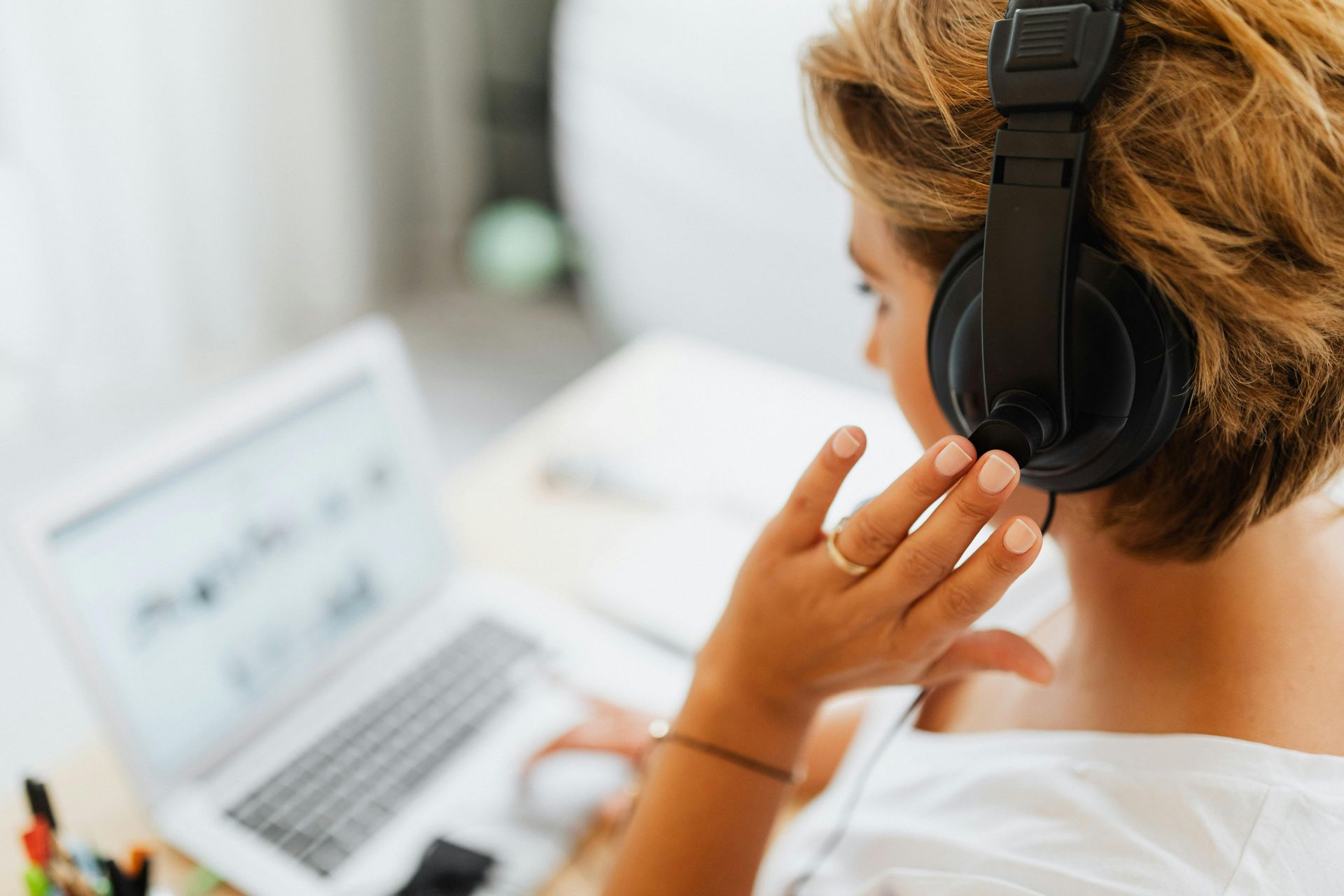 Woman wearing headphones, working on a laptop. Sitting indoors, she is wearing a white shirt and has a gold ring on her finger.