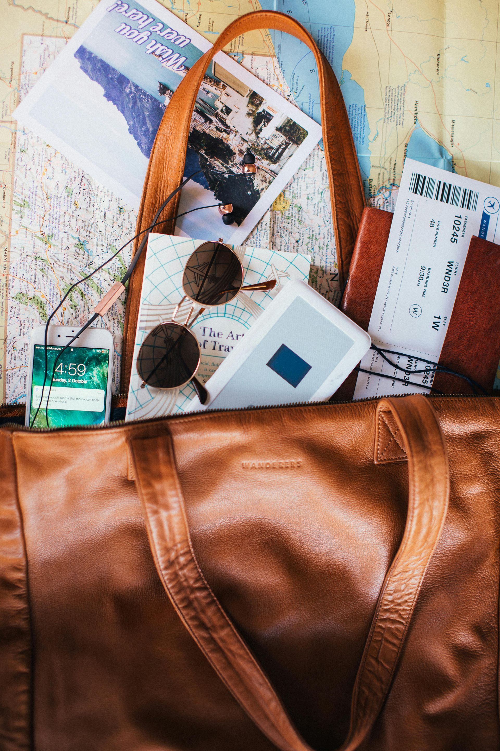 Brown leather travel bag overflowing with travel essentials, including maps, sunglasses, and a hand sanitizer.