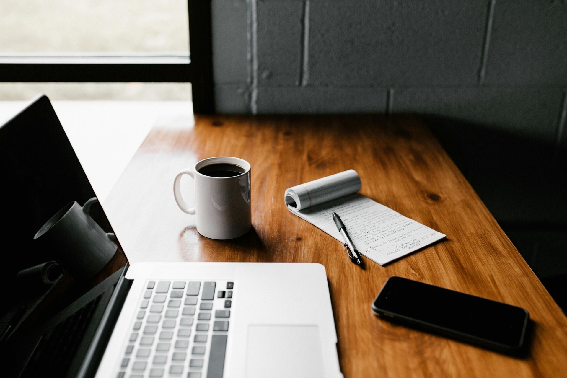 Laptop, coffee mug, notepad, pen, and phone on a wooden desk near a window.