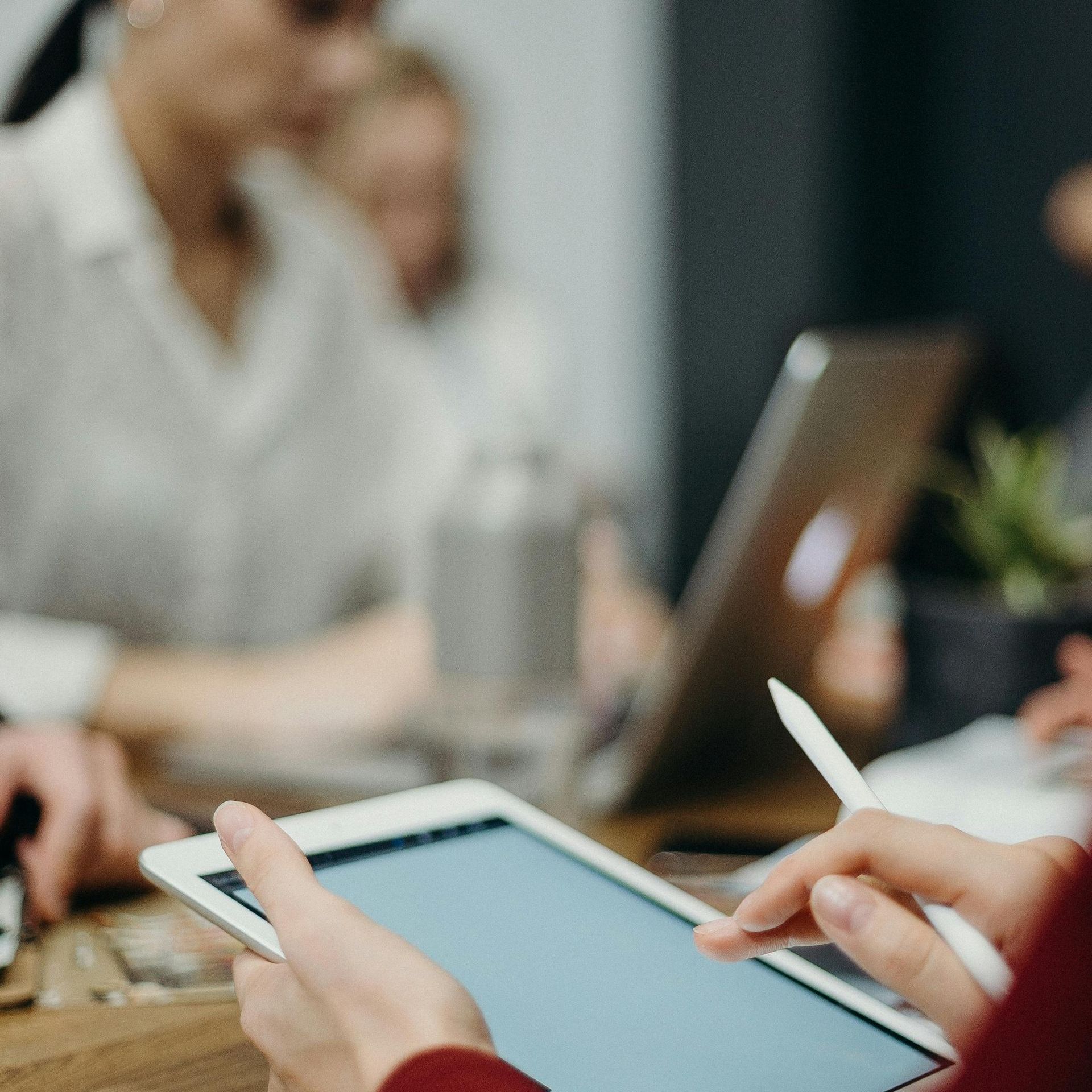 Person holding tablet with stylus at a table. Others work on laptops in a blurred office setting.