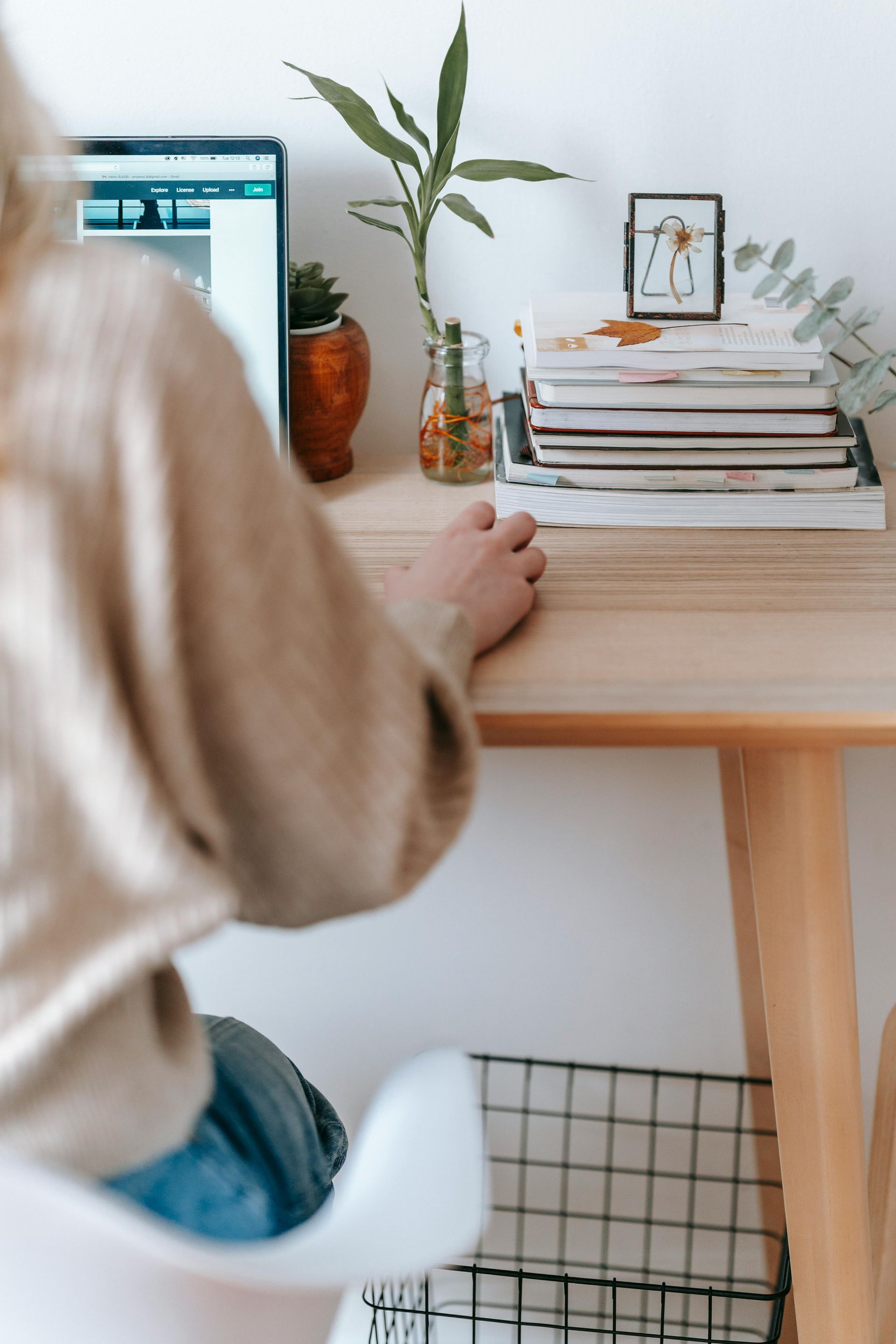 Person at a wooden desk with laptop, plants, and stack of books.