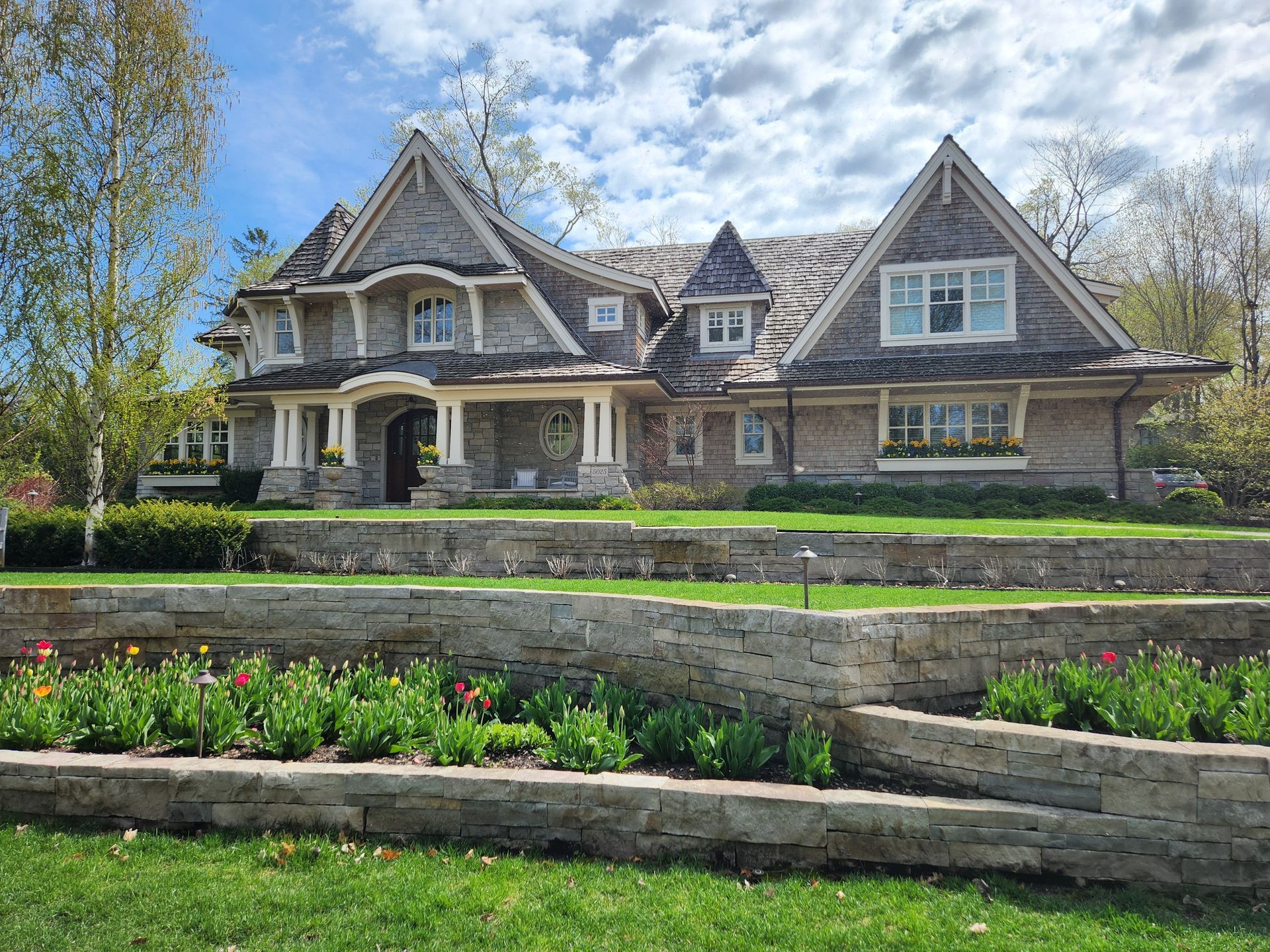 Large gray house with a stone retaining wall and flower beds, under a partly cloudy sky.