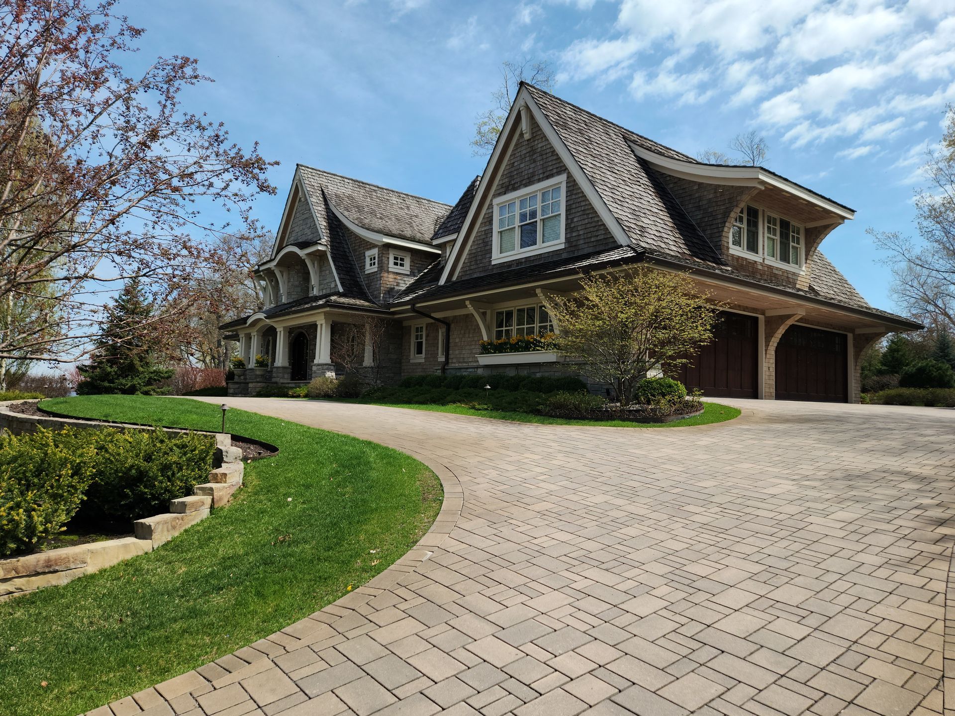House with brown shingle roof, stone exterior, and brick driveway. Green lawn and blue sky.