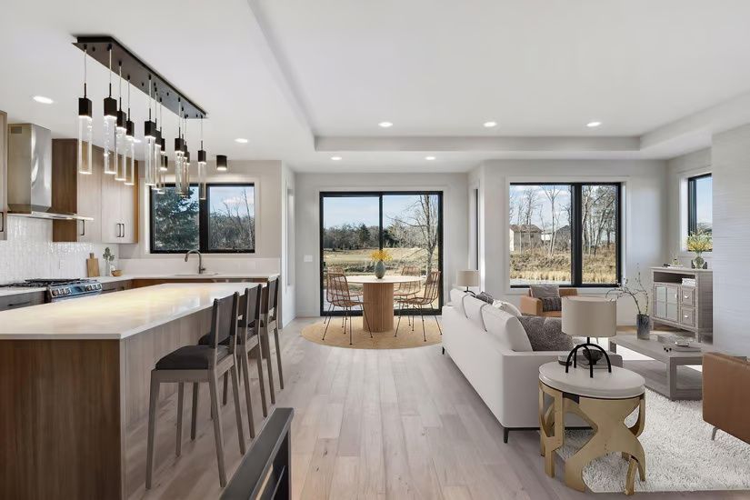 Open-concept kitchen and living area, light wood floors, white walls, large windows with outdoor views.