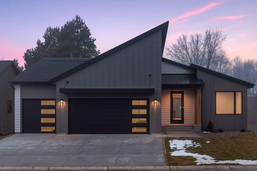 Modern gray house with angled roof, black garage doors, and a front door with sidelights; dusk setting.