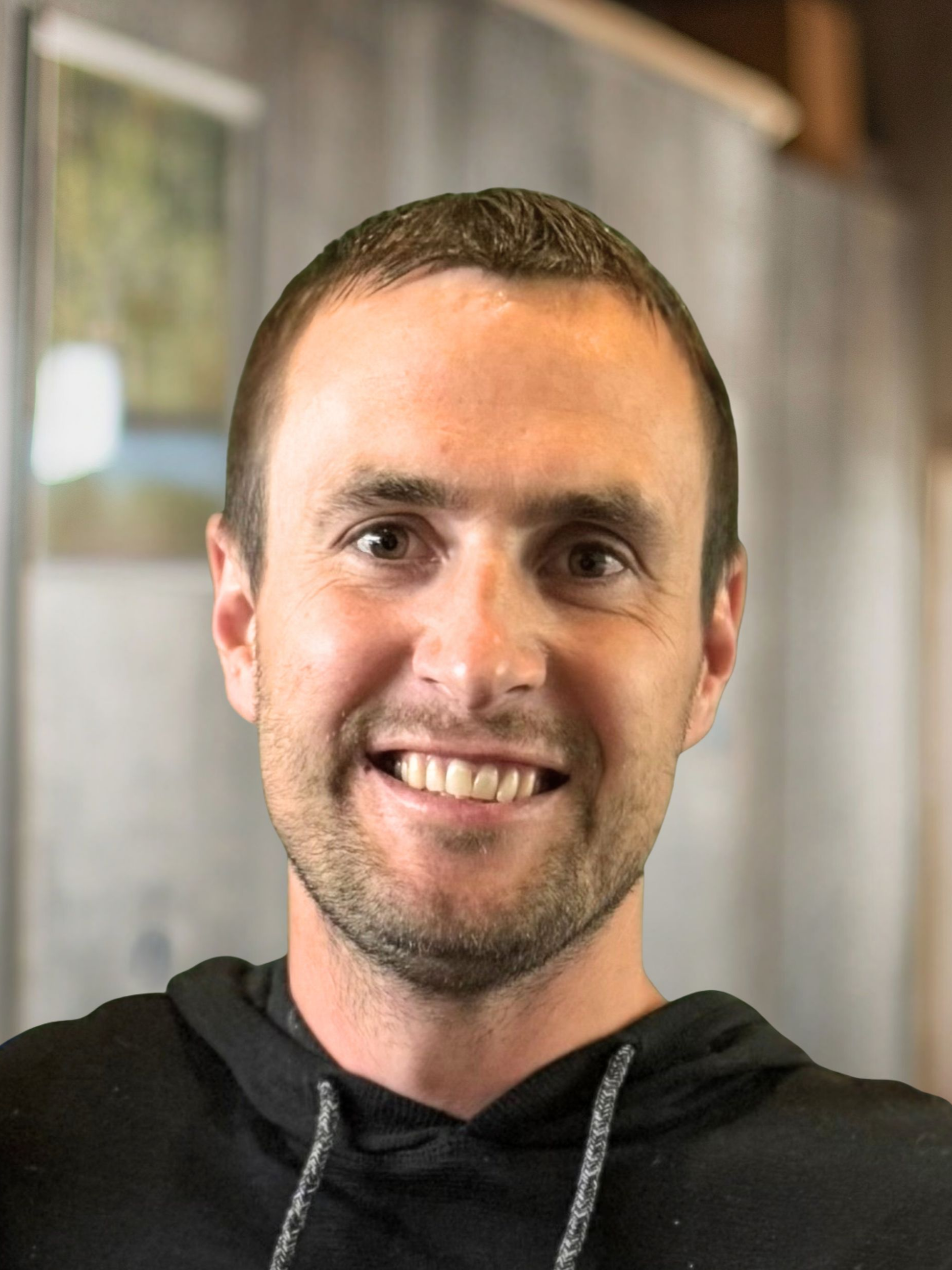 Man with short brown hair smiles, wearing a black hoodie in front of a neutral backdrop.