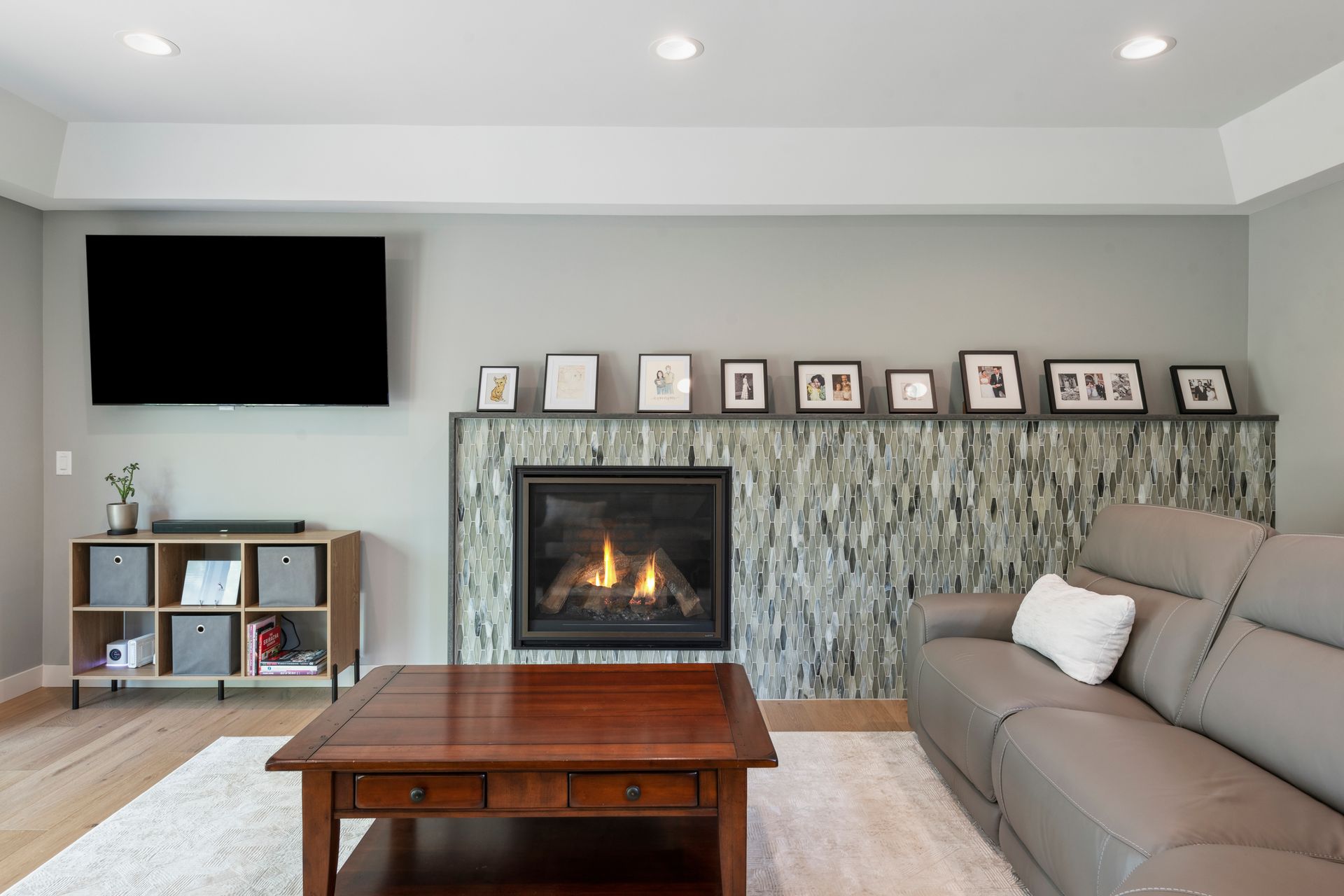 Living room with fireplace, TV, and gray leather sofa. Wooden furniture, light wood floor, gray walls.