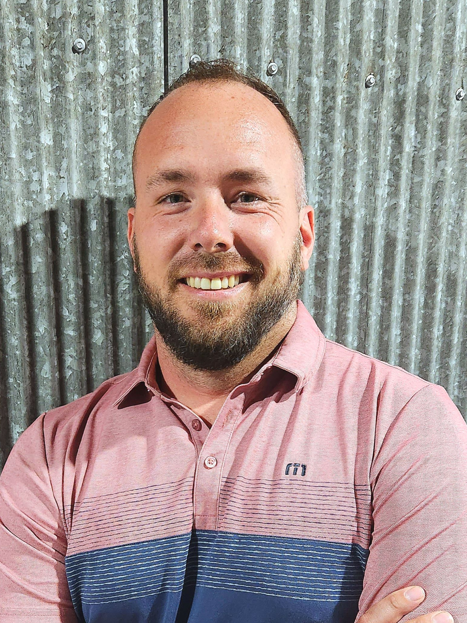 Man with a beard smiling, wearing a pink and blue striped shirt, arms crossed, standing in front of a corrugated metal wall.