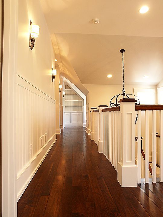 Hallway with dark wood floor, white walls, and a staircase. Overhead lighting and sconces illuminate the space.