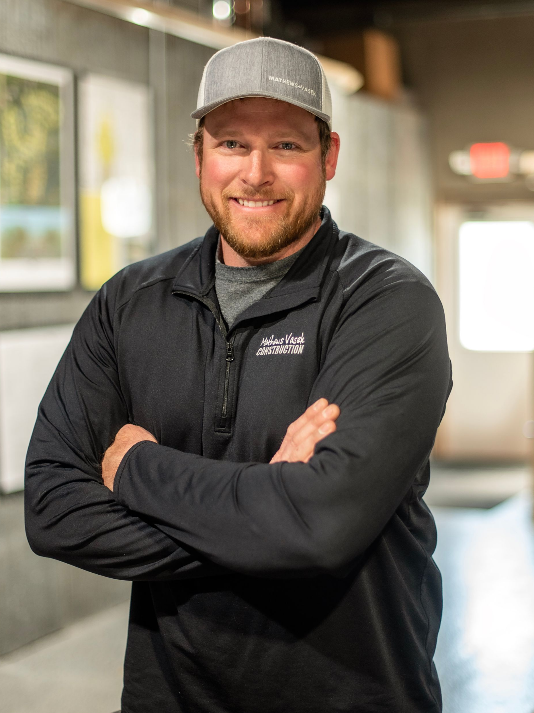 Man in black jacket and cap, arms crossed, smiling in hallway.
