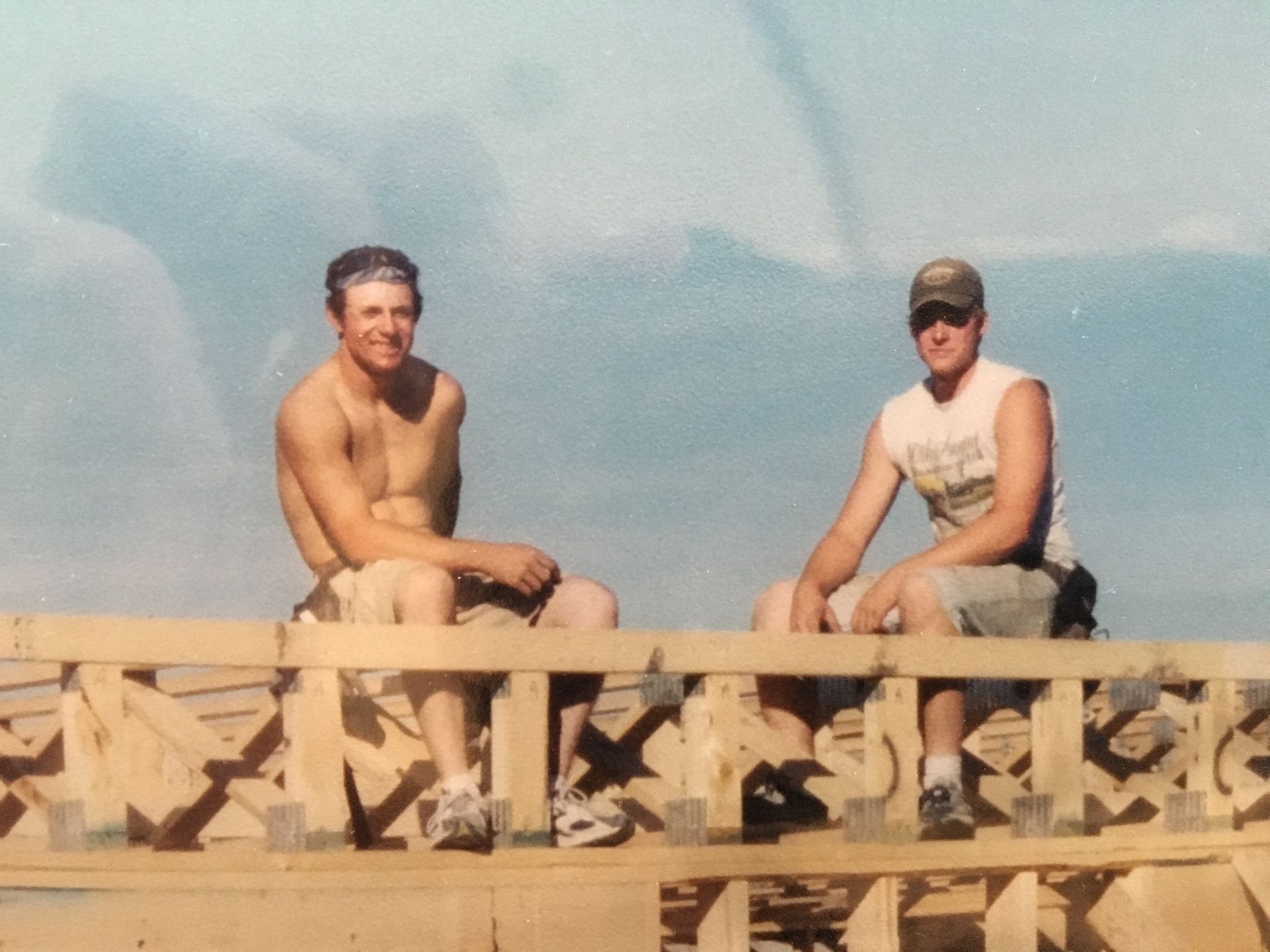 Two men sitting on wooden beams, one shirtless, the other in a tank top and cap; blue sky background.