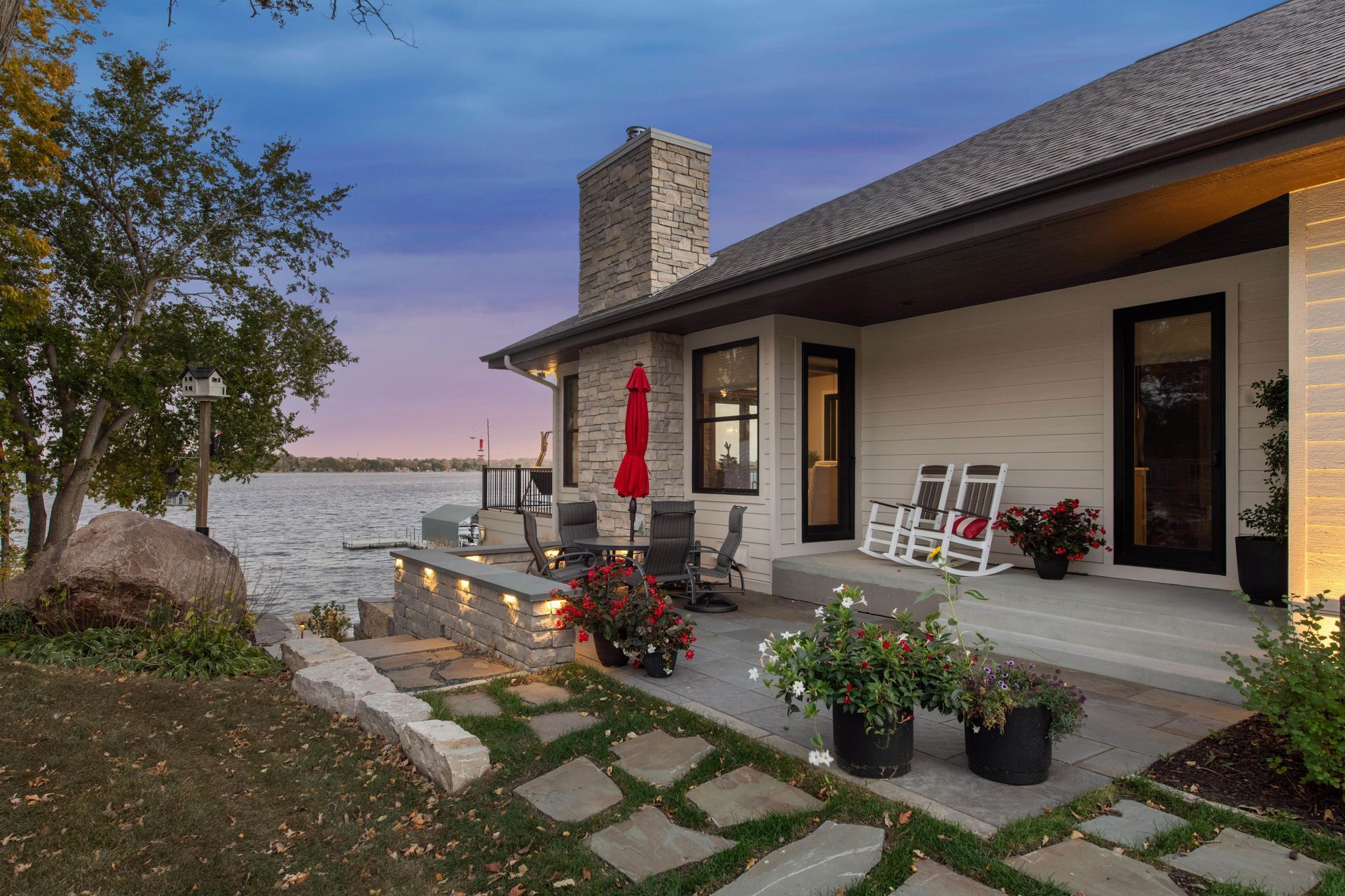House with patio overlooking water at dusk. Includes outdoor seating, landscaping, and brick chimney.
