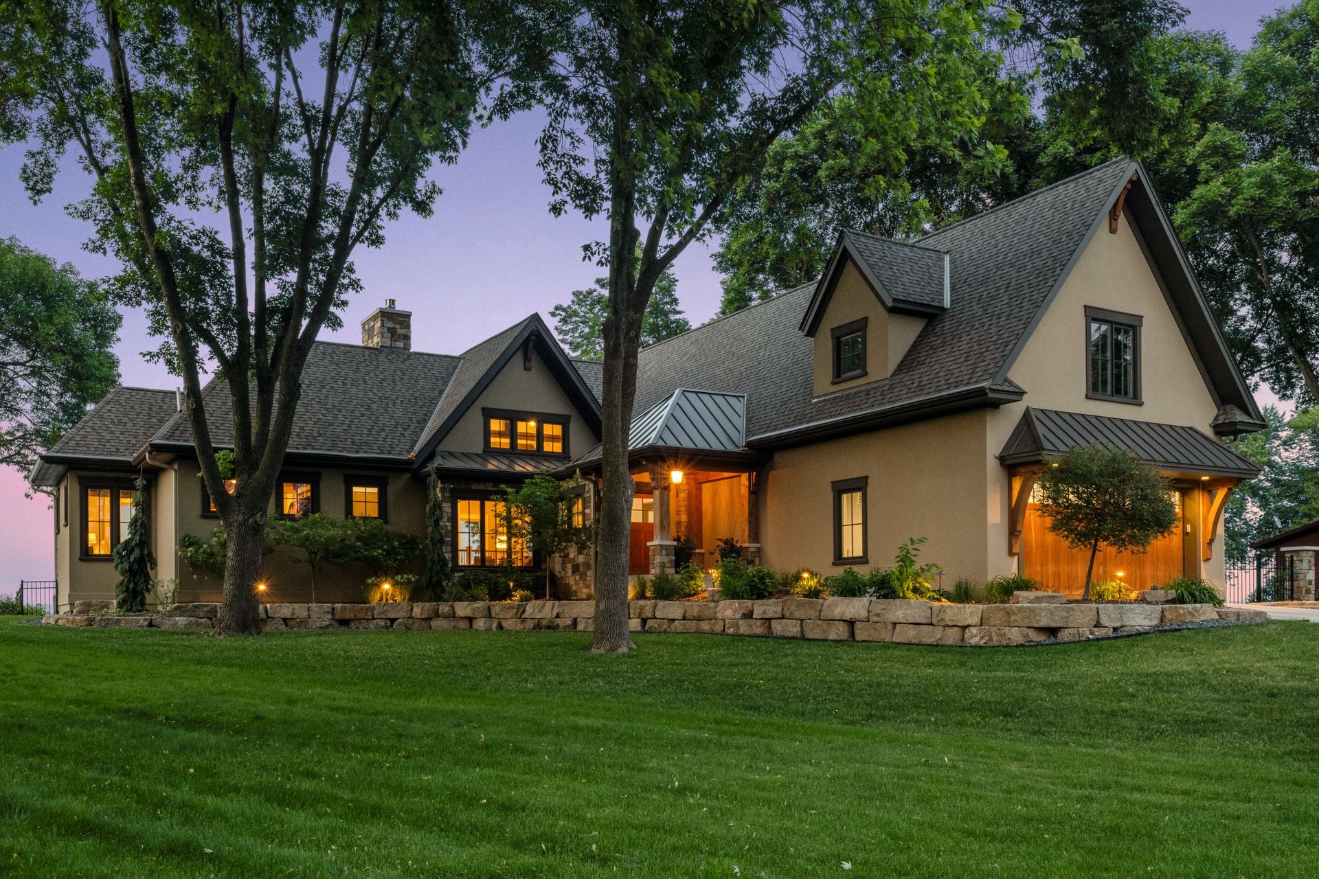 Tan house with dark roof and trim, lit windows, stone wall, and green lawn.