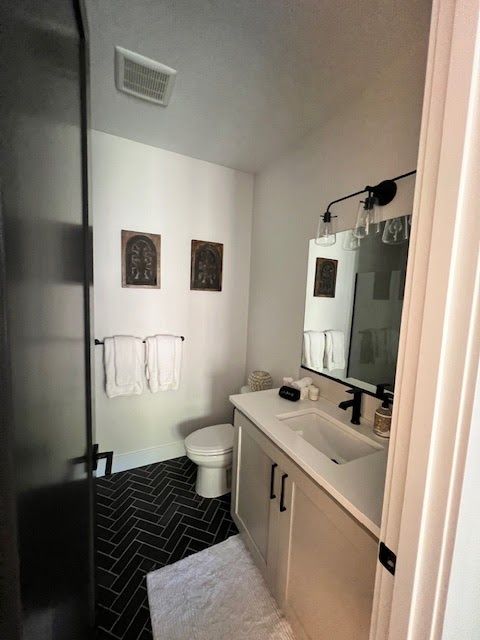 Small bathroom with white walls, black herringbone floor, white vanity, toilet, and towels.