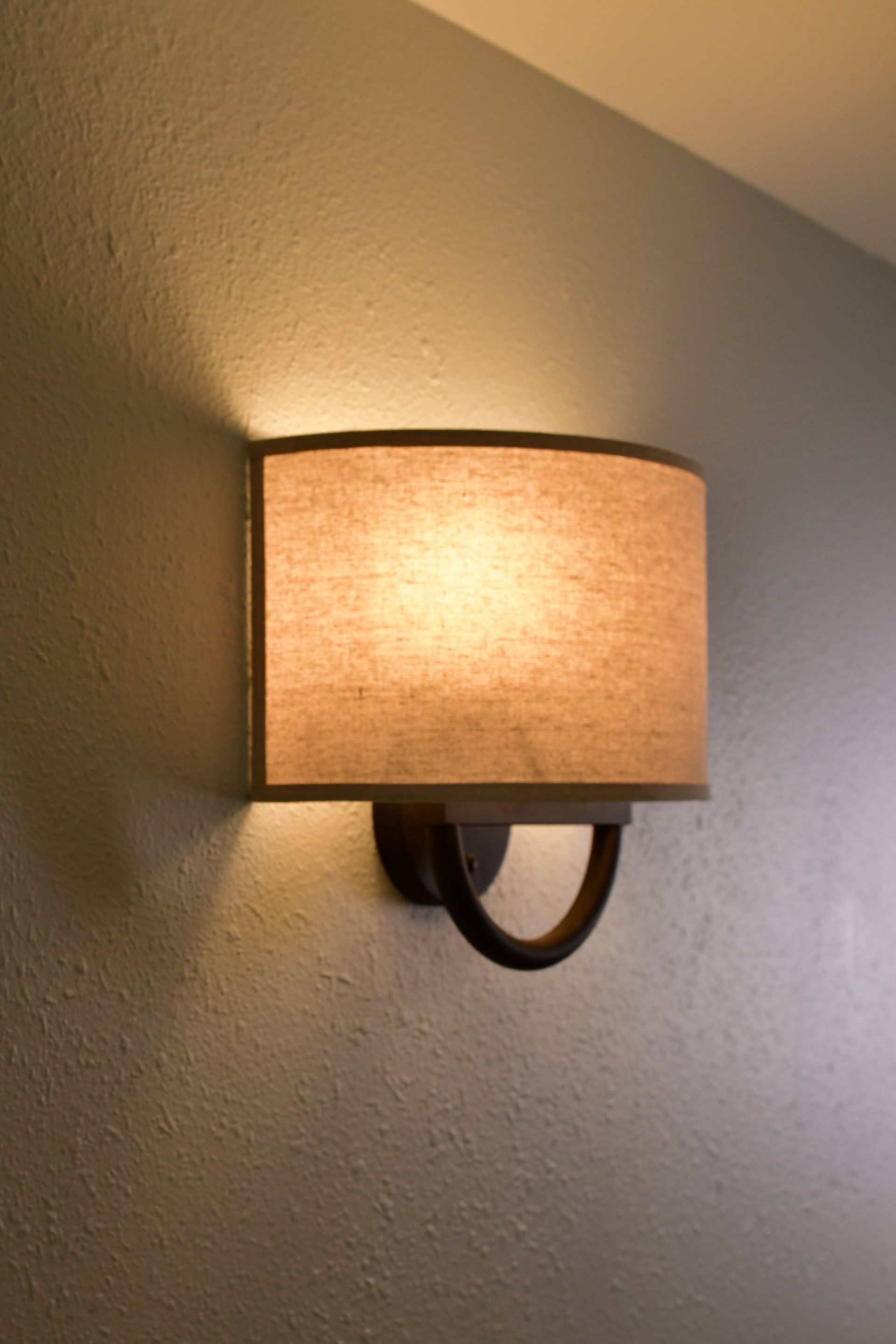 Wall-mounted lamp with a beige lampshade casting a warm glow on a textured wall.