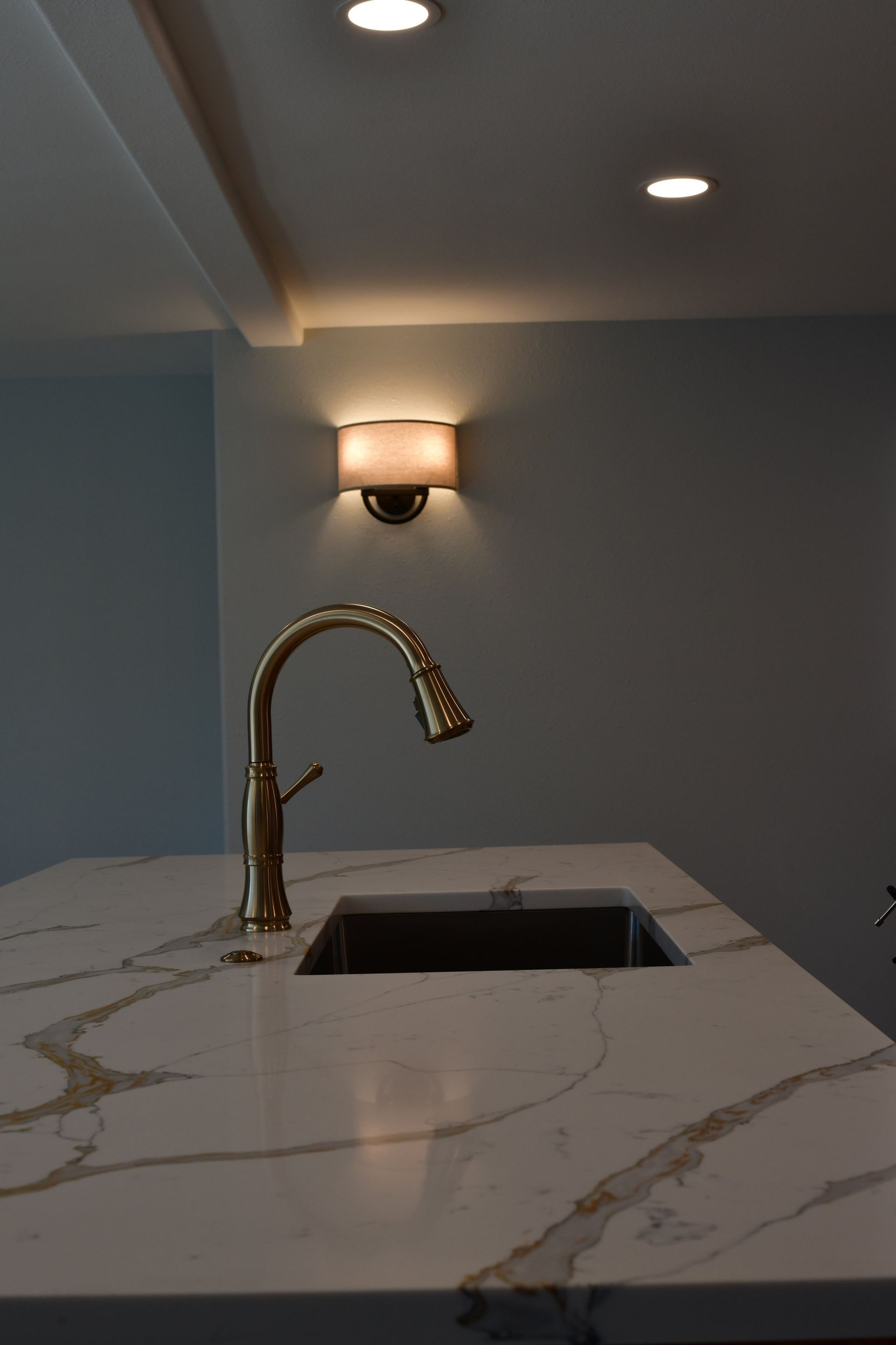 Kitchen island with gold faucet, dark sink, white countertop, and sconce on a gray wall.