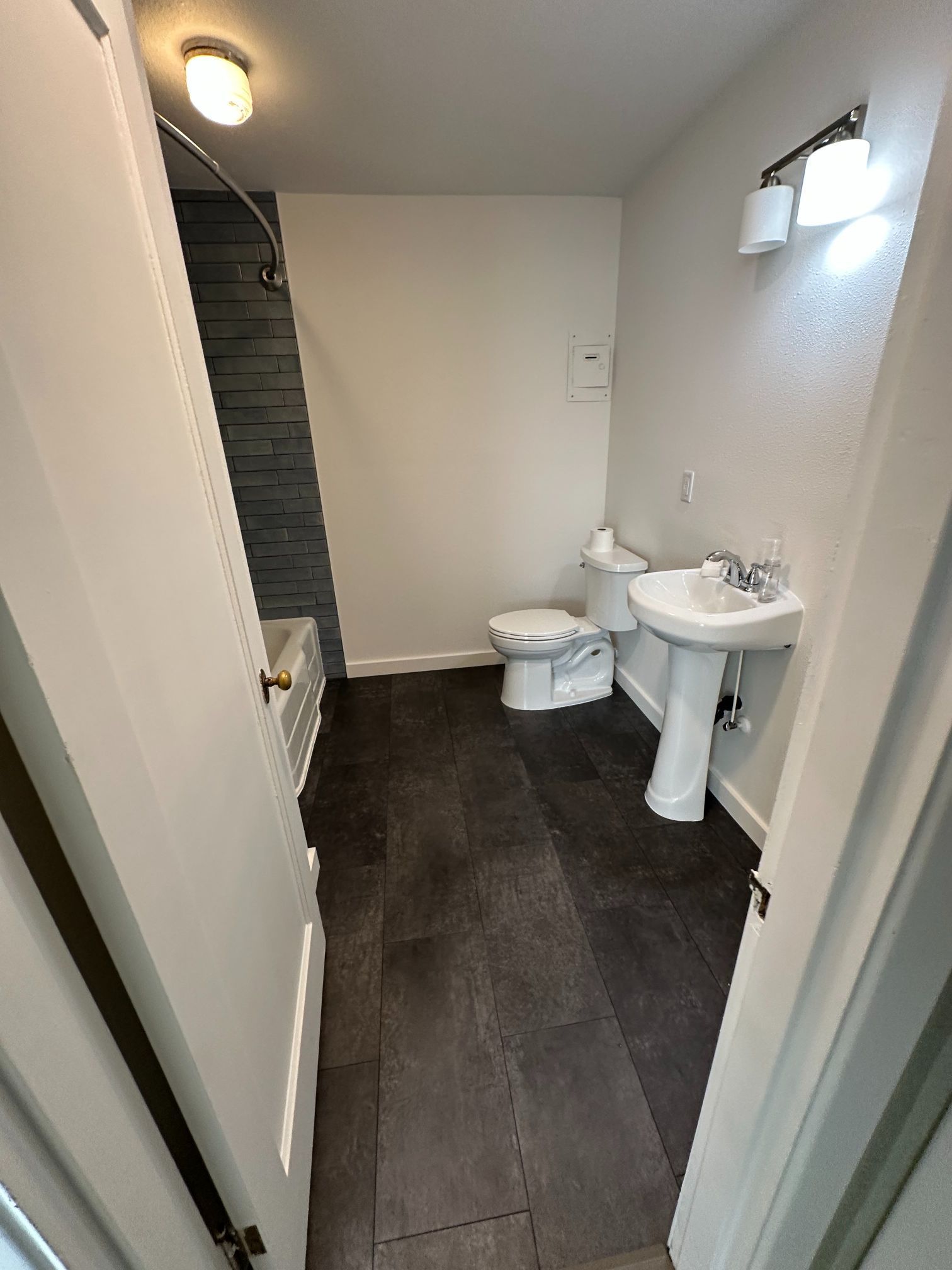 Small bathroom with white walls, dark floor, toilet, pedestal sink, and shower with gray tile.