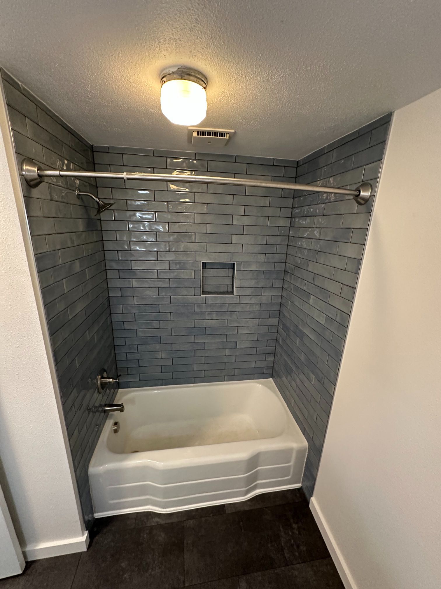 Blue tiled shower stall with a white tub, silver fixtures, and recessed shelf.