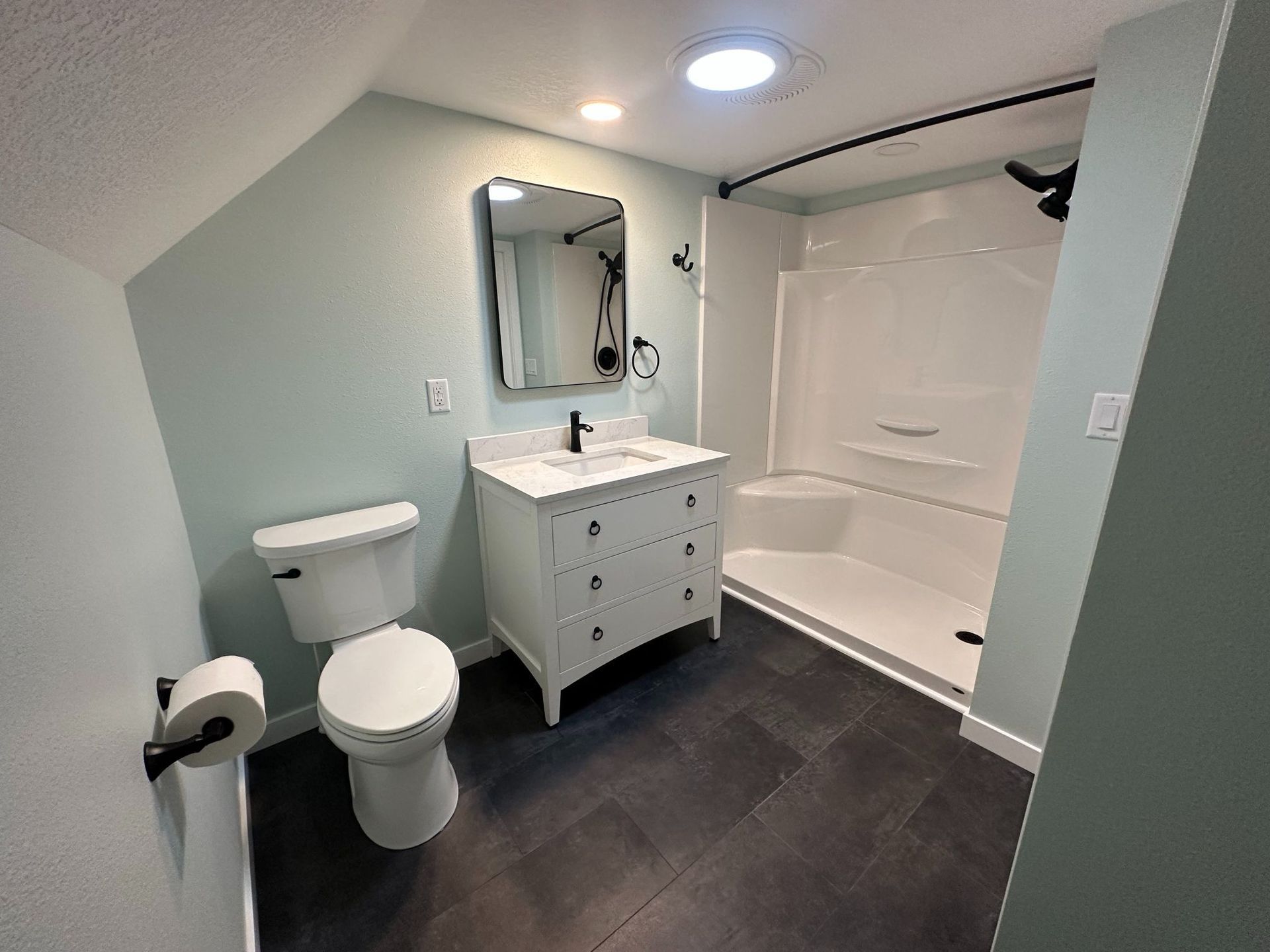 Bathroom with white vanity, toilet, and shower stall; light blue walls and dark gray tile floor.