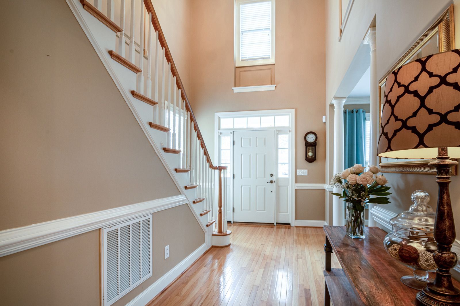 Entryway with wooden staircase, beige walls, white door, and table with lamp and flowers.