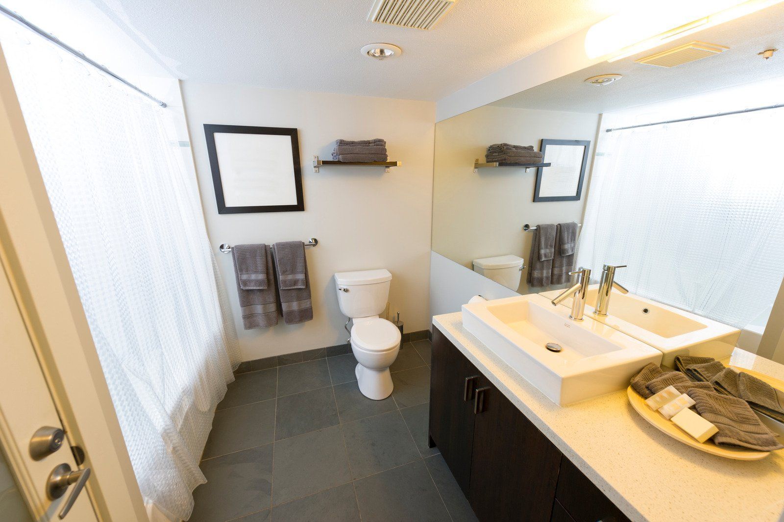 Bathroom with white sink, dark cabinets, toilet, large mirror, and gray tiled floor.