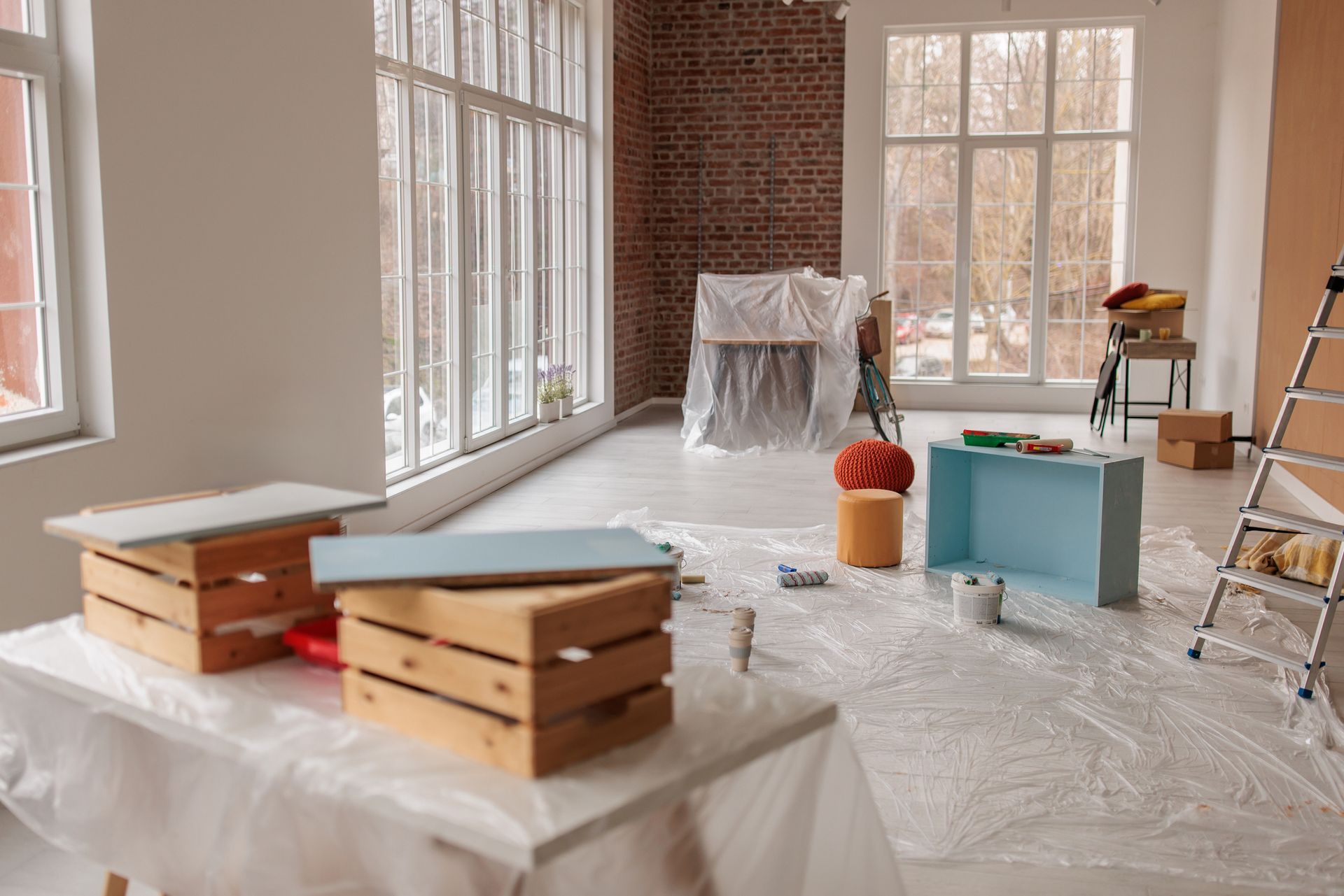 An interior room under renovation with wooden crates, a step ladder, drop cloths, and furniture covered in plastic.