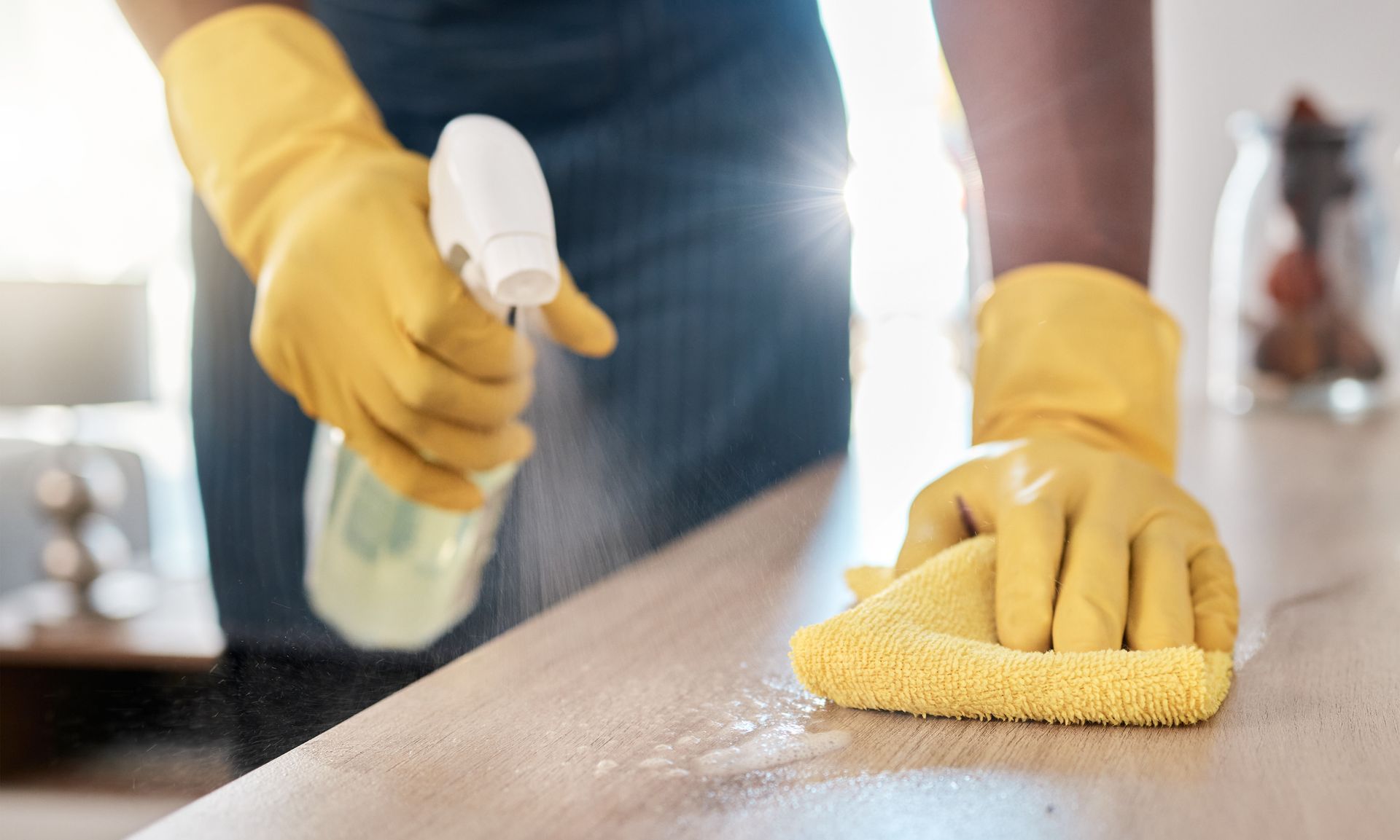 A person wearing yellow gloves is cleaning a counter with a cloth and spray bottle.