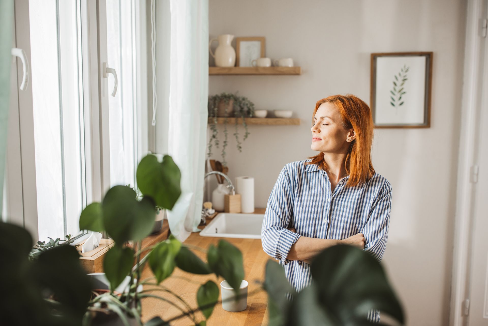 Woman with red hair looks out a window, arms crossed, in a bright kitchen.