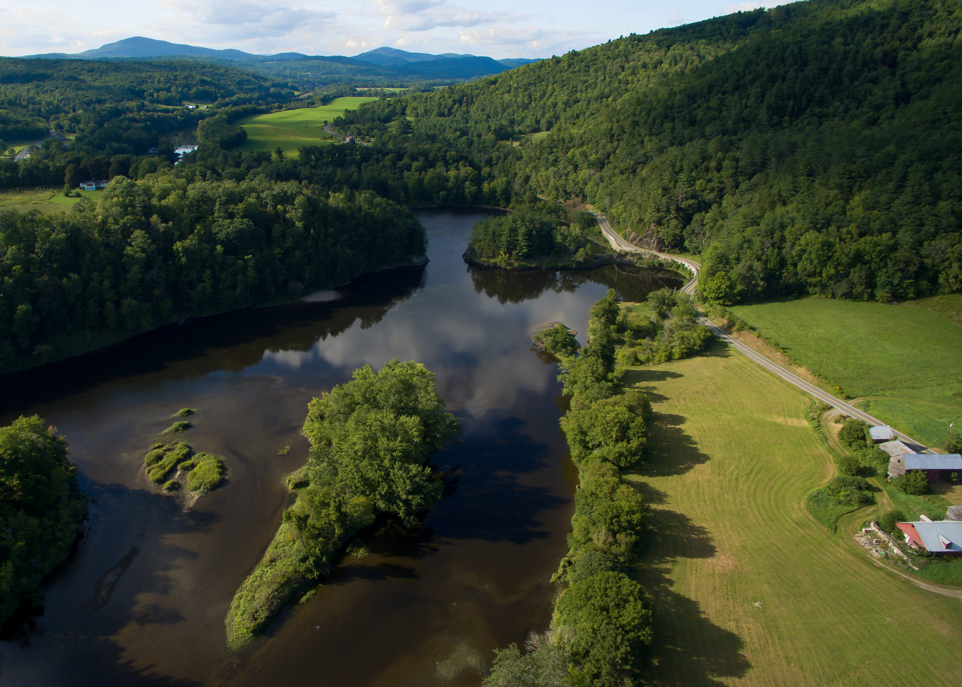 Aerial view of a river winding through a lush green valley, flanked by trees and a sunny meadow.