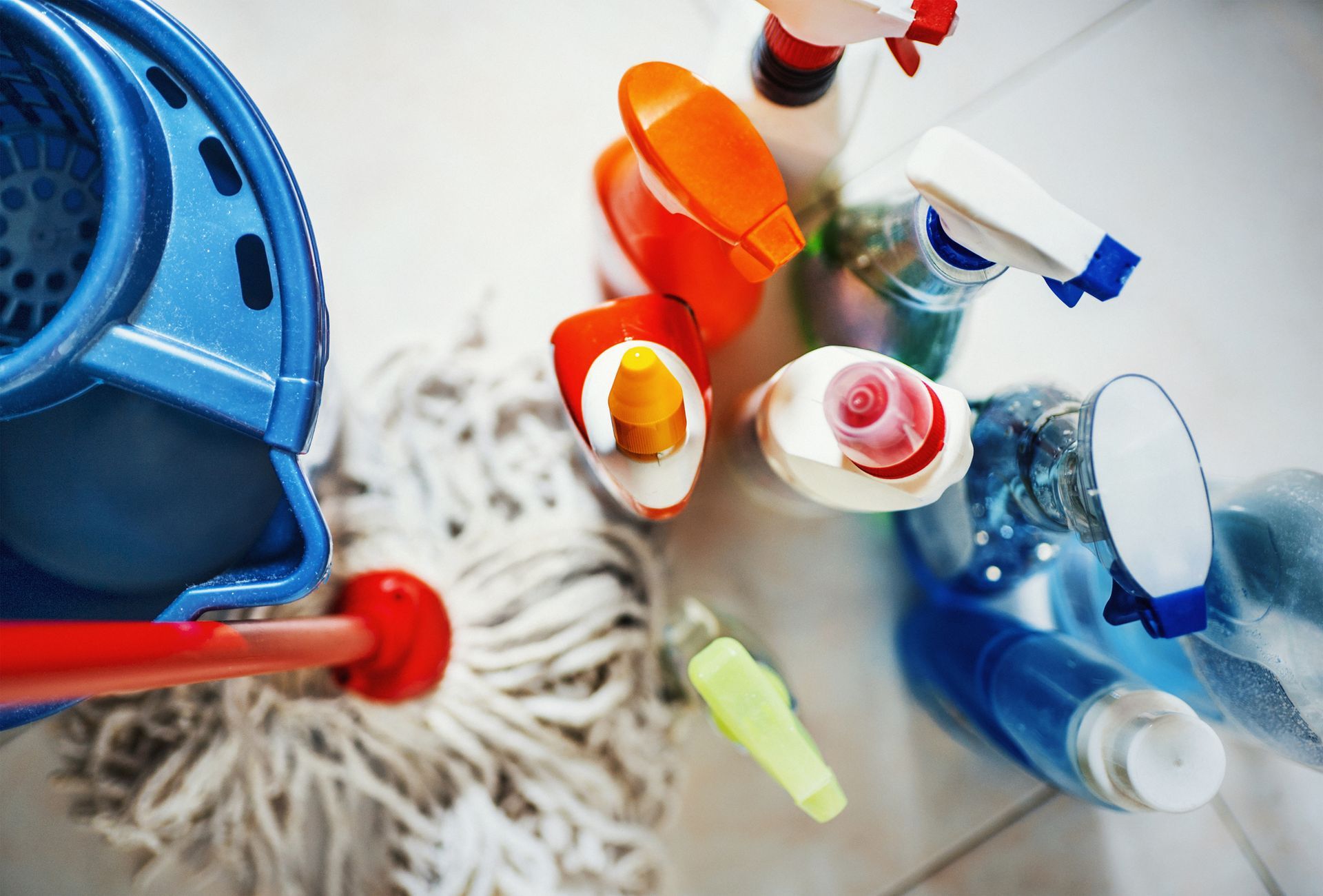 Blue mop and bucket with cleaning supplies on a white tile floor, overhead view.