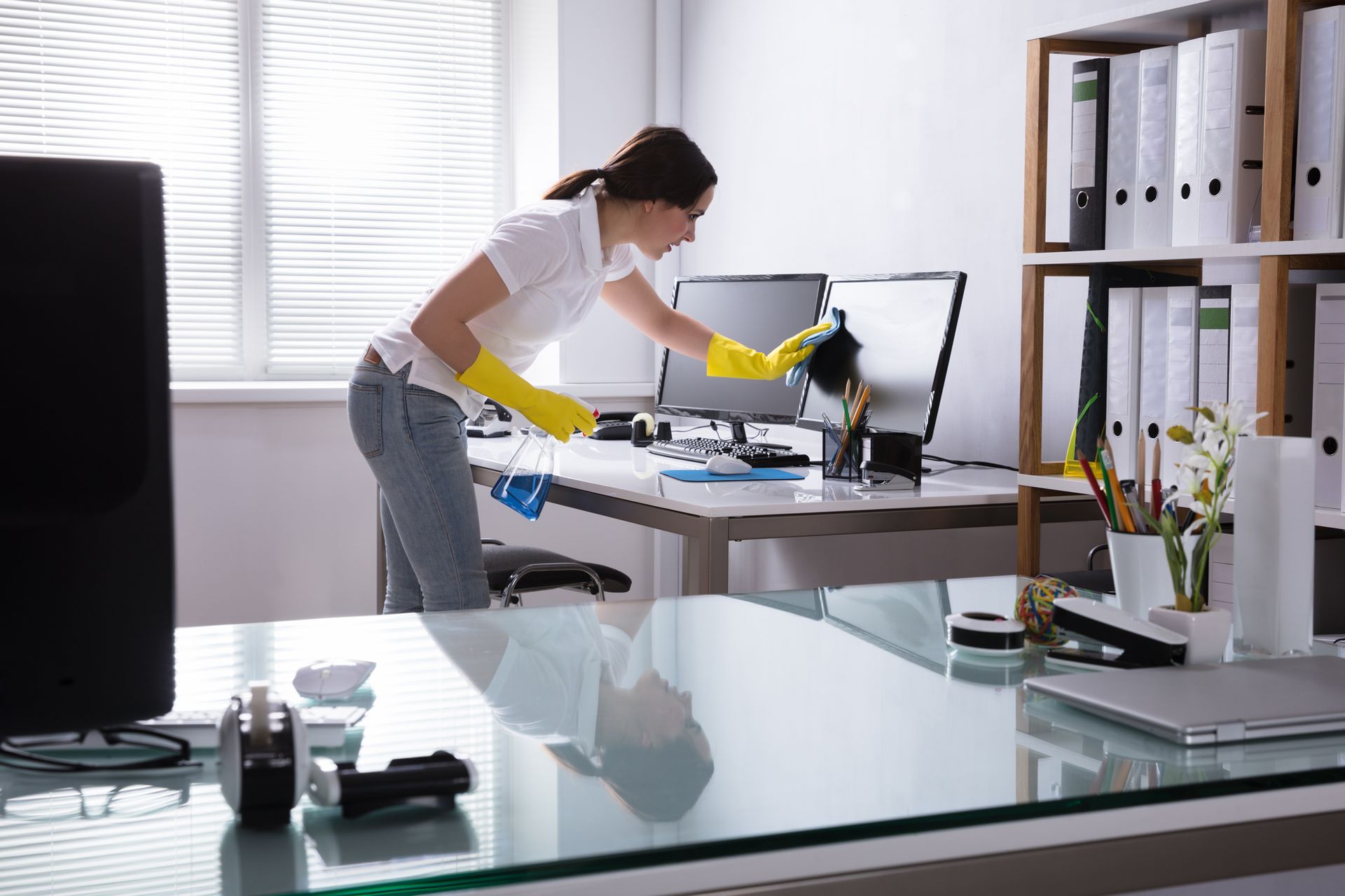 A woman is cleaning a desk in an office with a spray bottle.