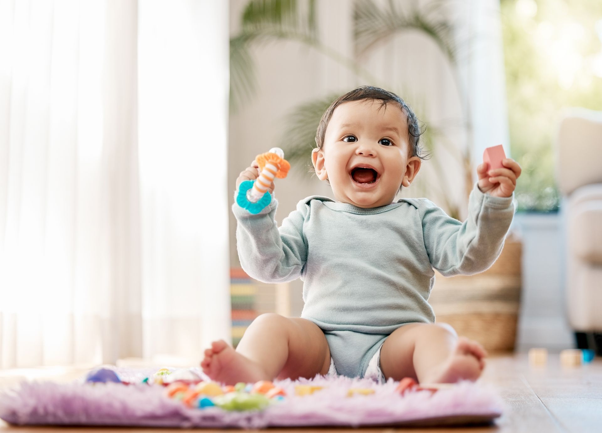 A baby sitting on a blanket with toys. The baby's mouth is open and hands are in the air.